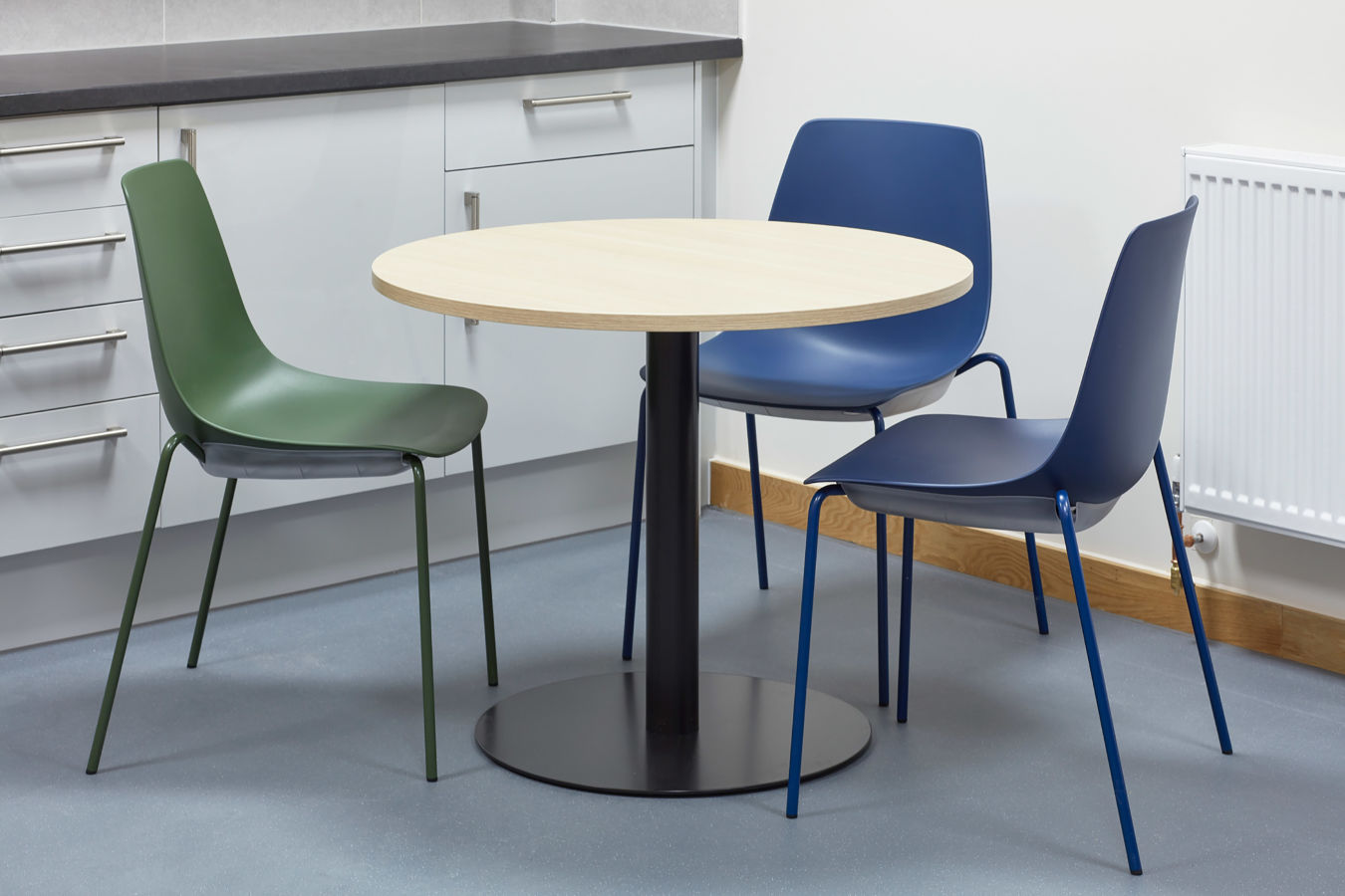 A round wooden table is surrounded by two blue chairs and a green chair in a modern kitchen with white cabinets and a radiator.