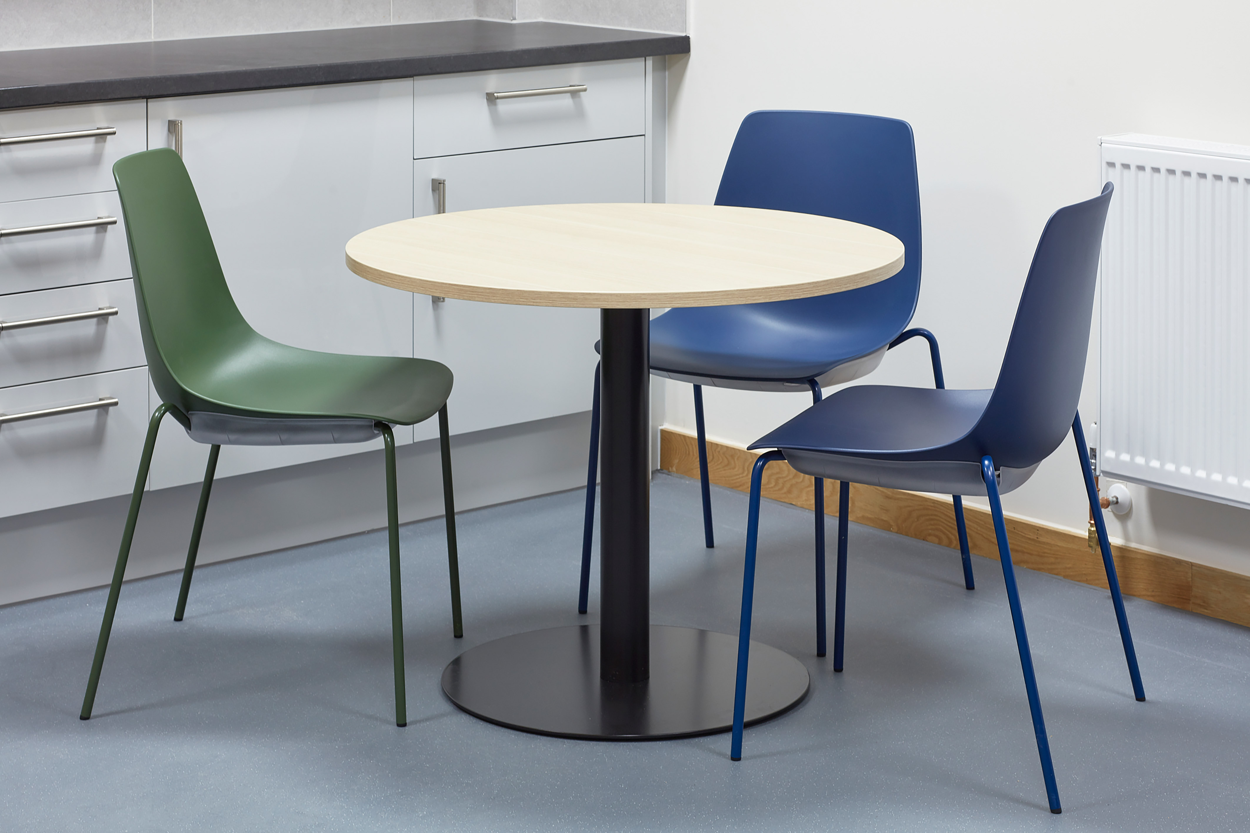 A round wooden table is surrounded by two blue chairs and a green chair in a modern kitchen with white cabinets and a radiator.