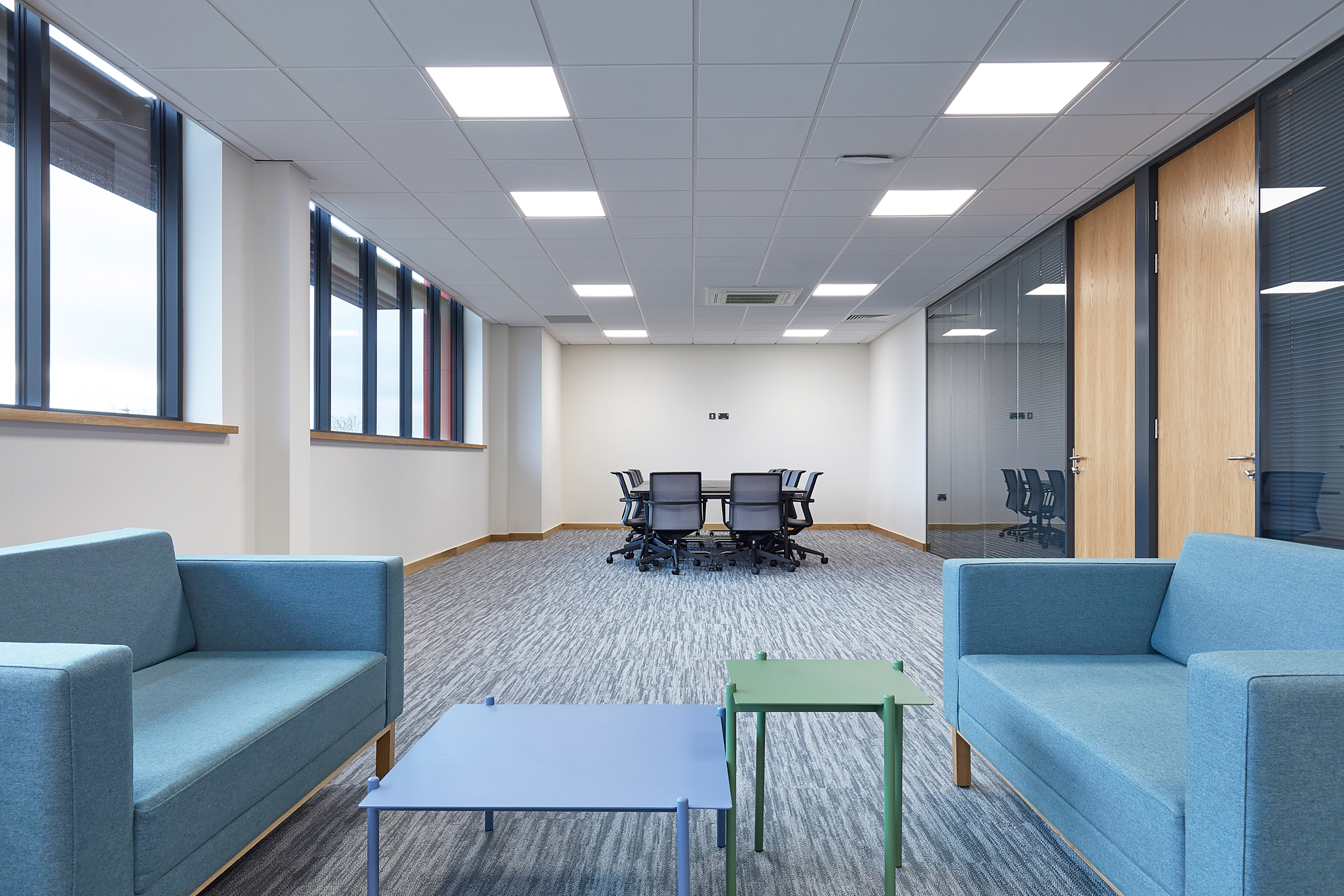 A modern office space features blue sofas and colorful tables in the foreground, with a conference table and chairs in the background. Spacious layout and large windows provide natural light.