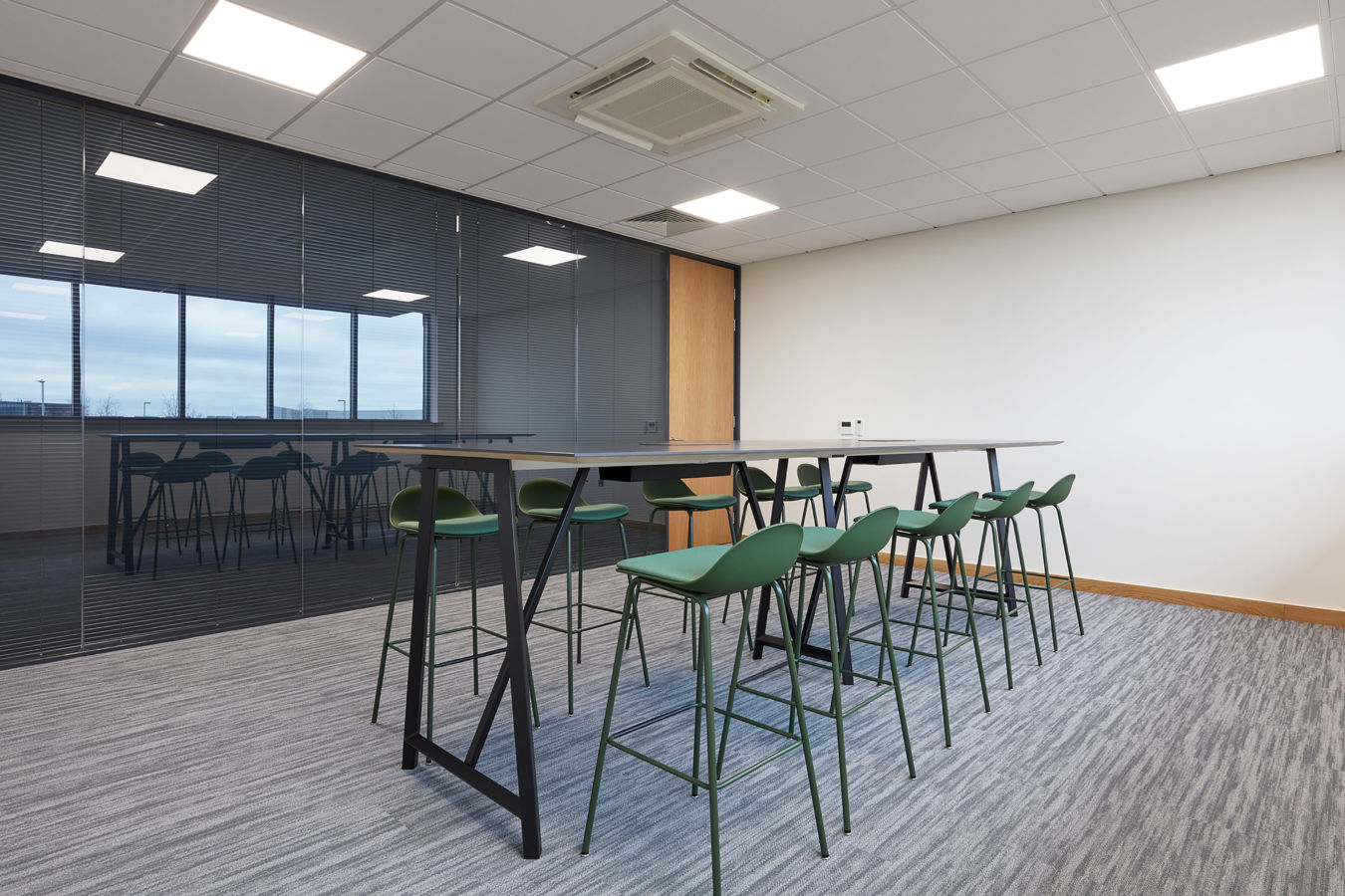 modern conference room with a tall black table and green high stools on a gray carpet. Glass wall, bright lighting, and minimalist design convey a professional tone