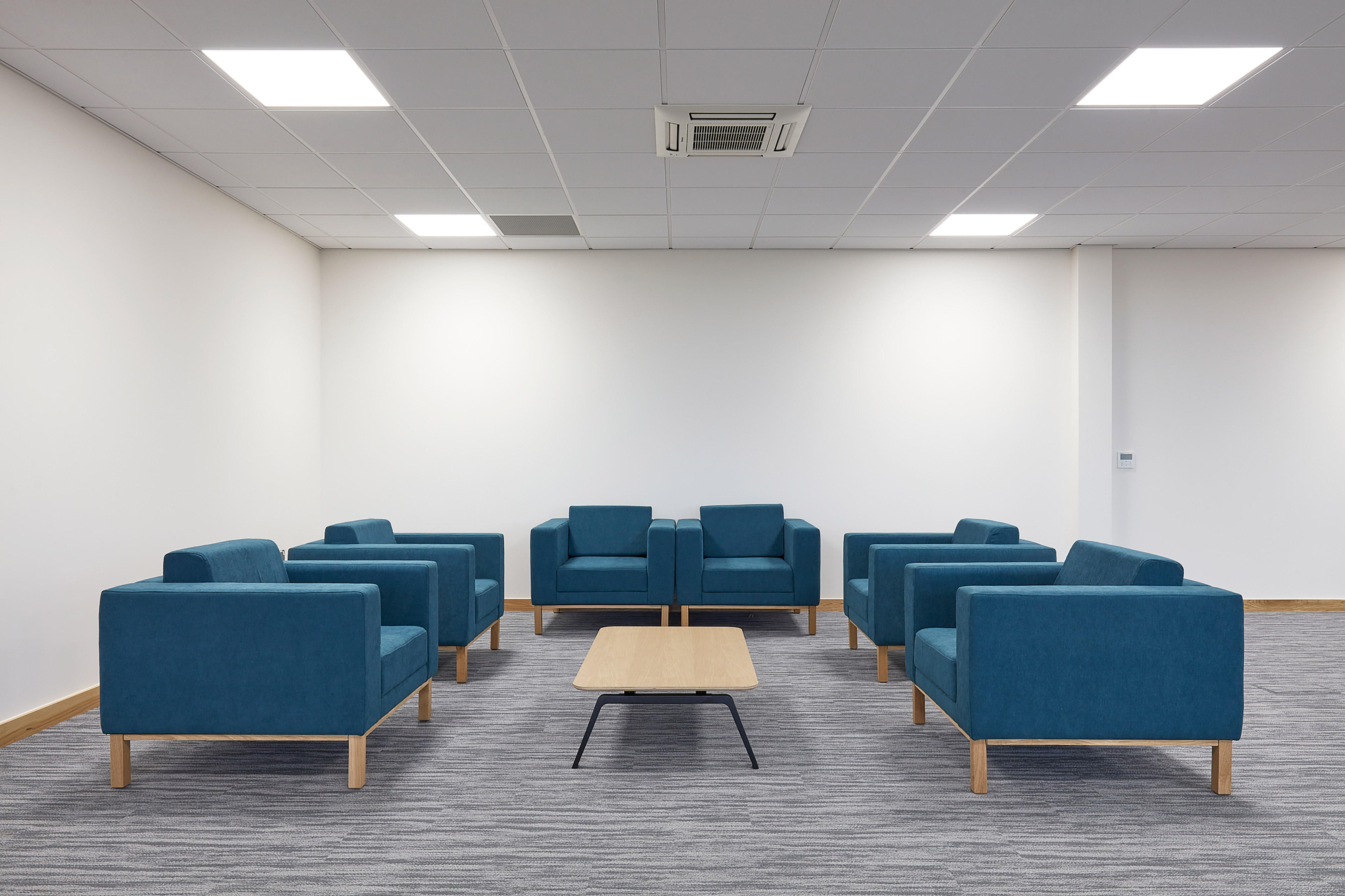 Blue armchairs encircle a small wooden coffee table, situated on a gray carpet in a minimalist, brightly lit room with white walls and a tiled ceiling.