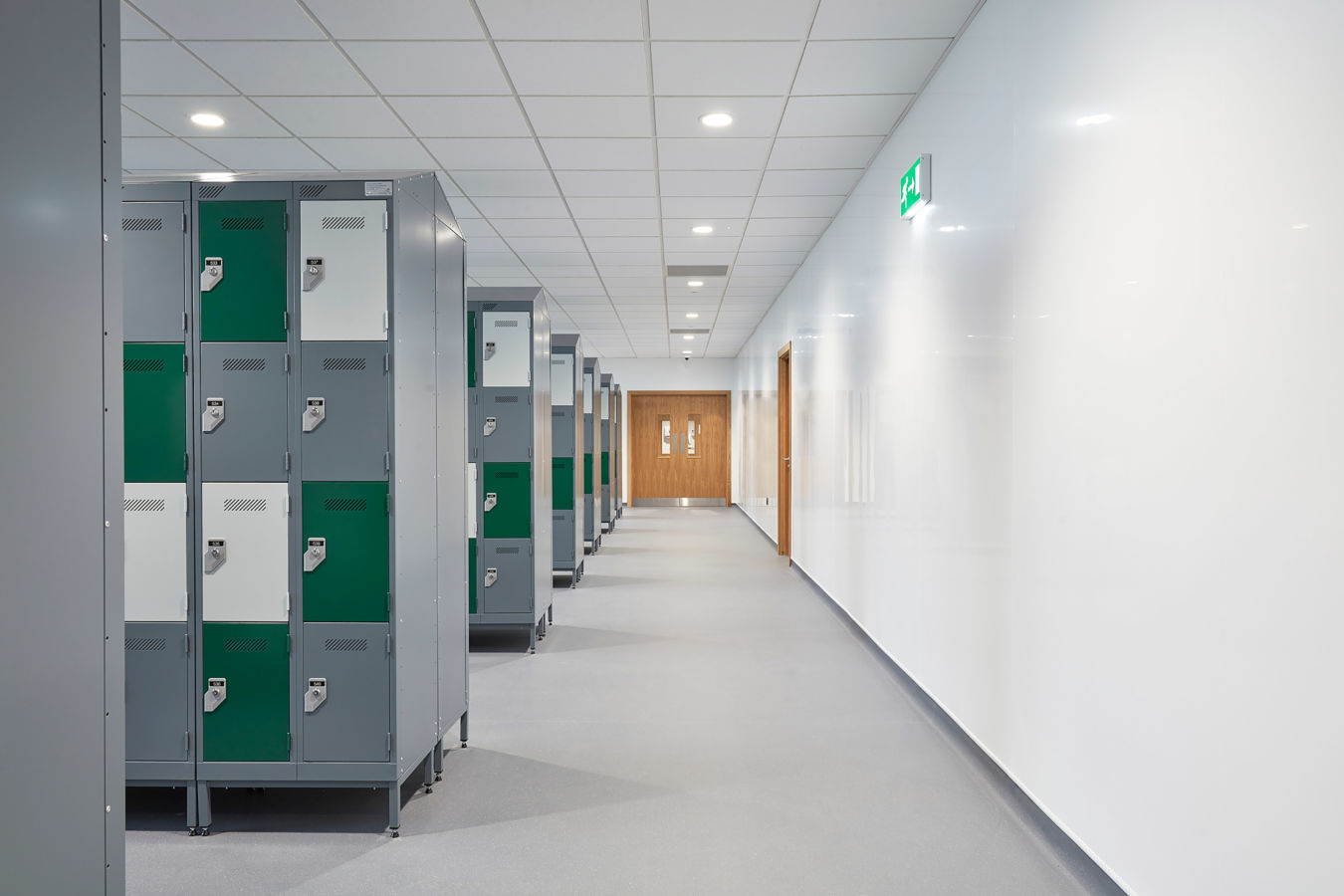 clean, modern hallway with rows of gray and green lockers on the left. White walls and ceiling lead to wooden doors, creating a bright, orderly atmosphere