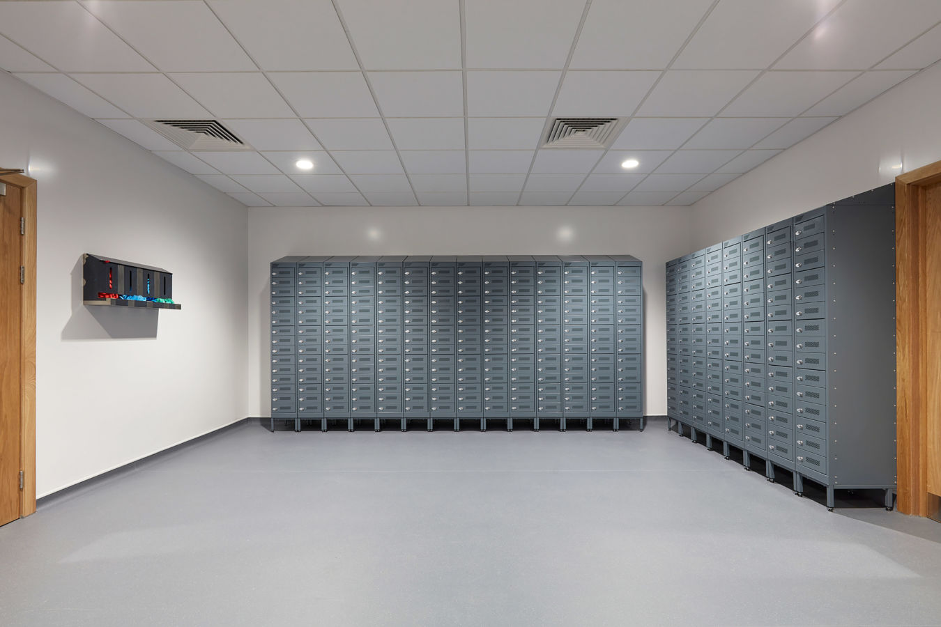 clean, bright room with gray lockers covering two walls. A small rack with colorful items hangs on the left wall. The space feels organized and modern