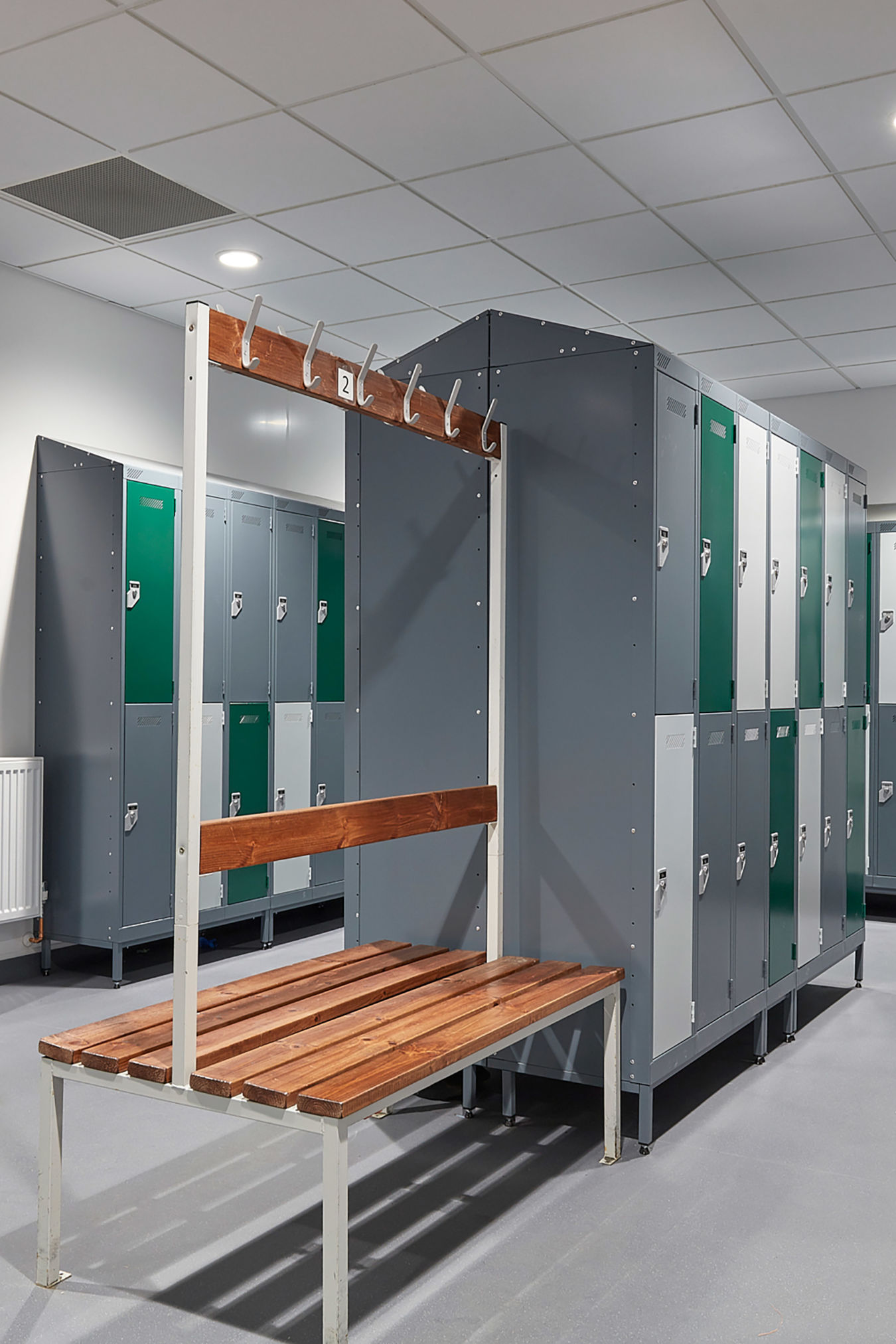 Modern locker room with gray and green lockers, wooden benches, and hooks. Clean, well-lit space with tiled ceiling, evoking a tidy, organized atmosphere