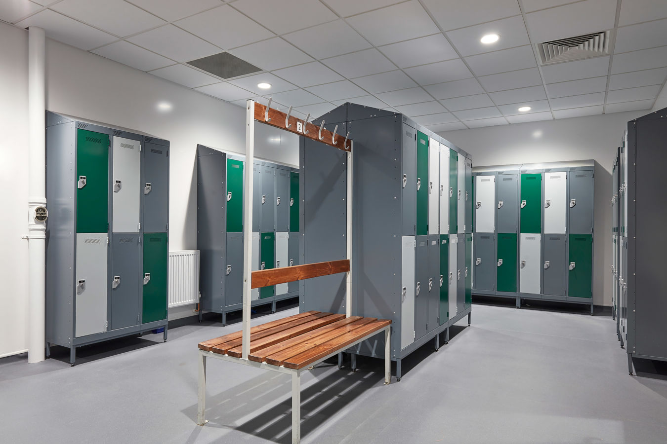Modern locker room with gray and green lockers, wooden benches, and hooks. Clean, well-lit space with tiled ceiling, evoking a tidy, organized atmosphere