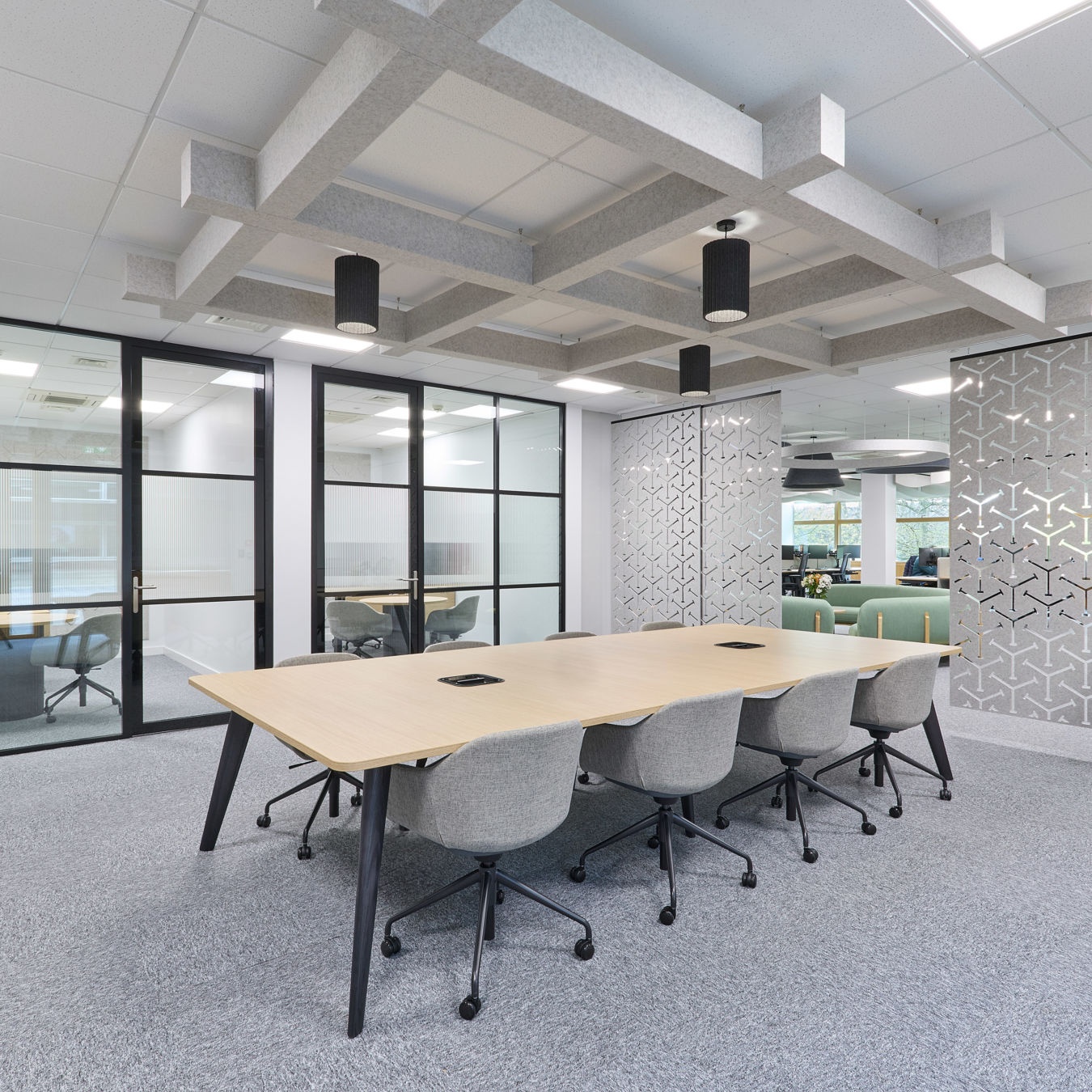 A conference table with eight grey chairs is arranged in a modern office, surrounded by glass partitions and geometric-patterned walls, with suspended ceiling decor and natural light through windows.