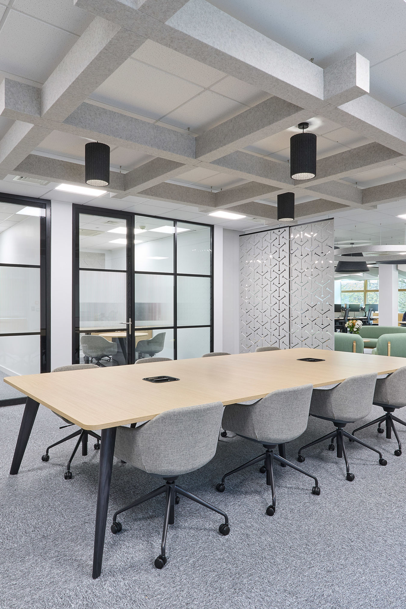 A conference table with eight grey chairs is arranged in a modern office, surrounded by glass partitions and geometric-patterned walls, with suspended ceiling decor and natural light through windows.