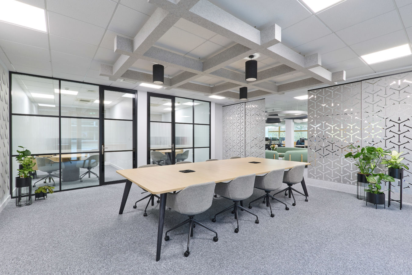 A conference table with eight grey chairs is arranged in a modern office, surrounded by glass partitions and geometric-patterned walls, with suspended ceiling decor and natural light through windows.