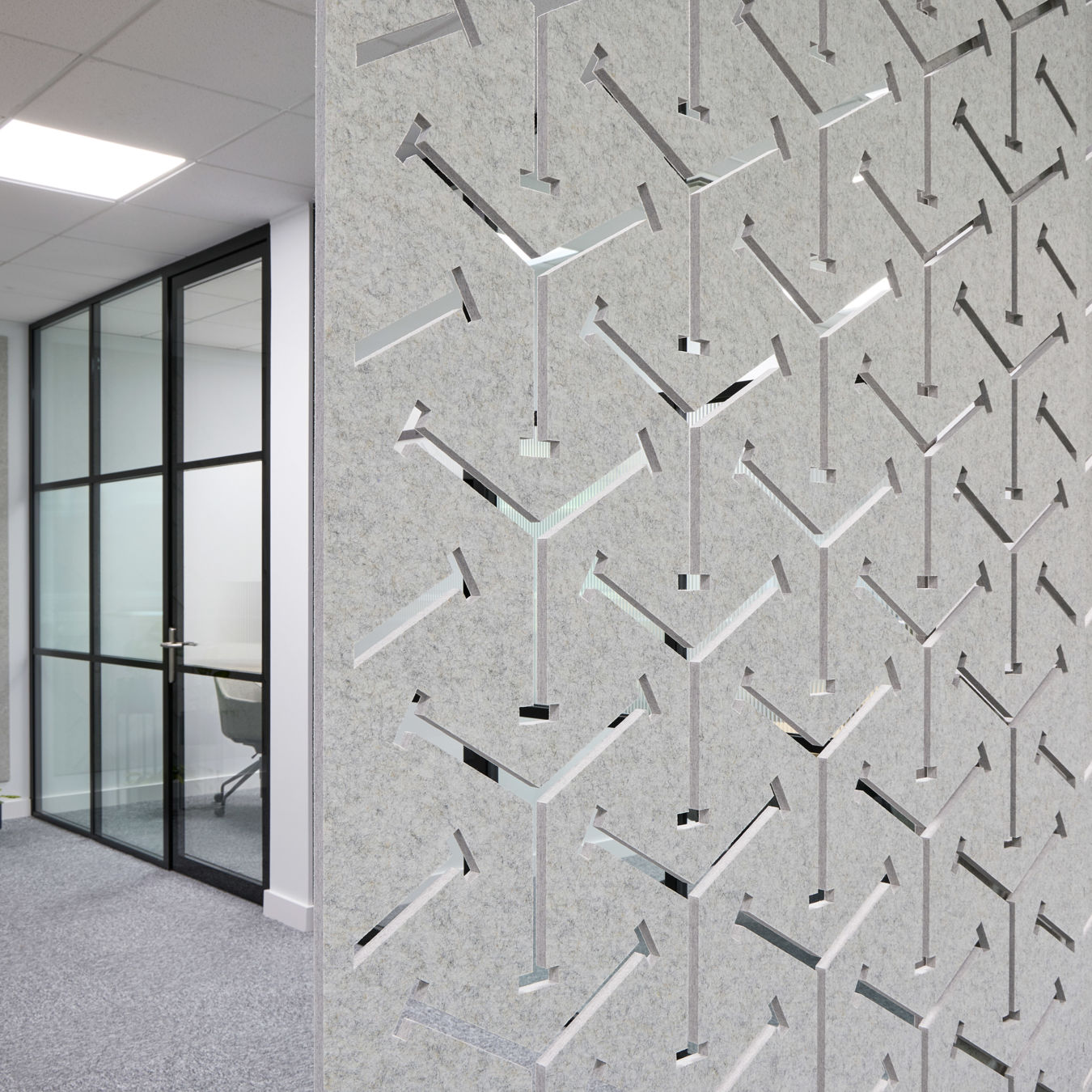 Decorative wall with geometric, arrow-shaped cutouts enhances an office space; glass partition door and indoor plants accentuate the modern, minimalist design.
