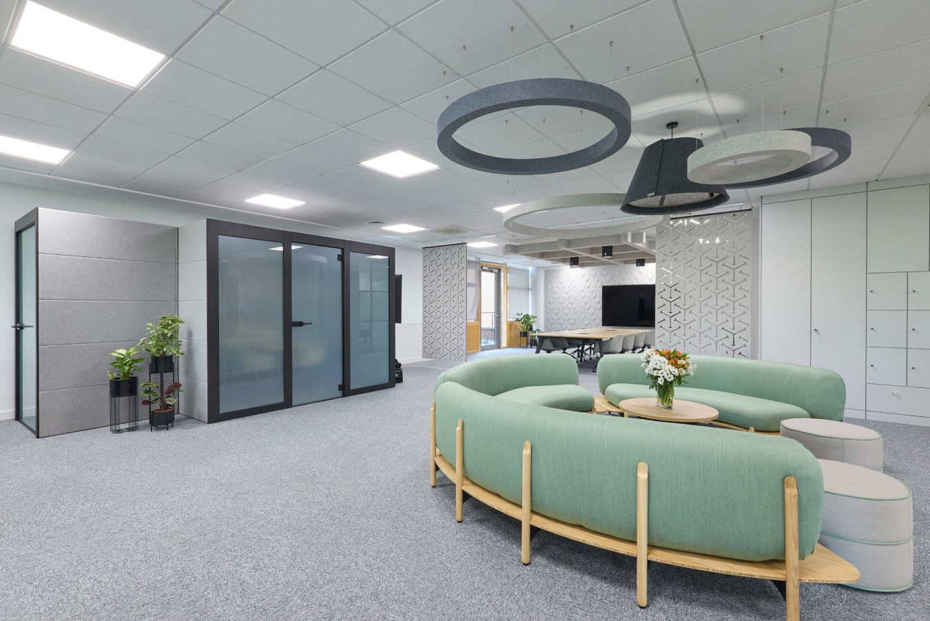 Modern office lounge features circular green sofas, coffee tables with flowers, and overhead ring lighting. Glass-walled meeting rooms and abstract wall designs create an open, stylish environment.