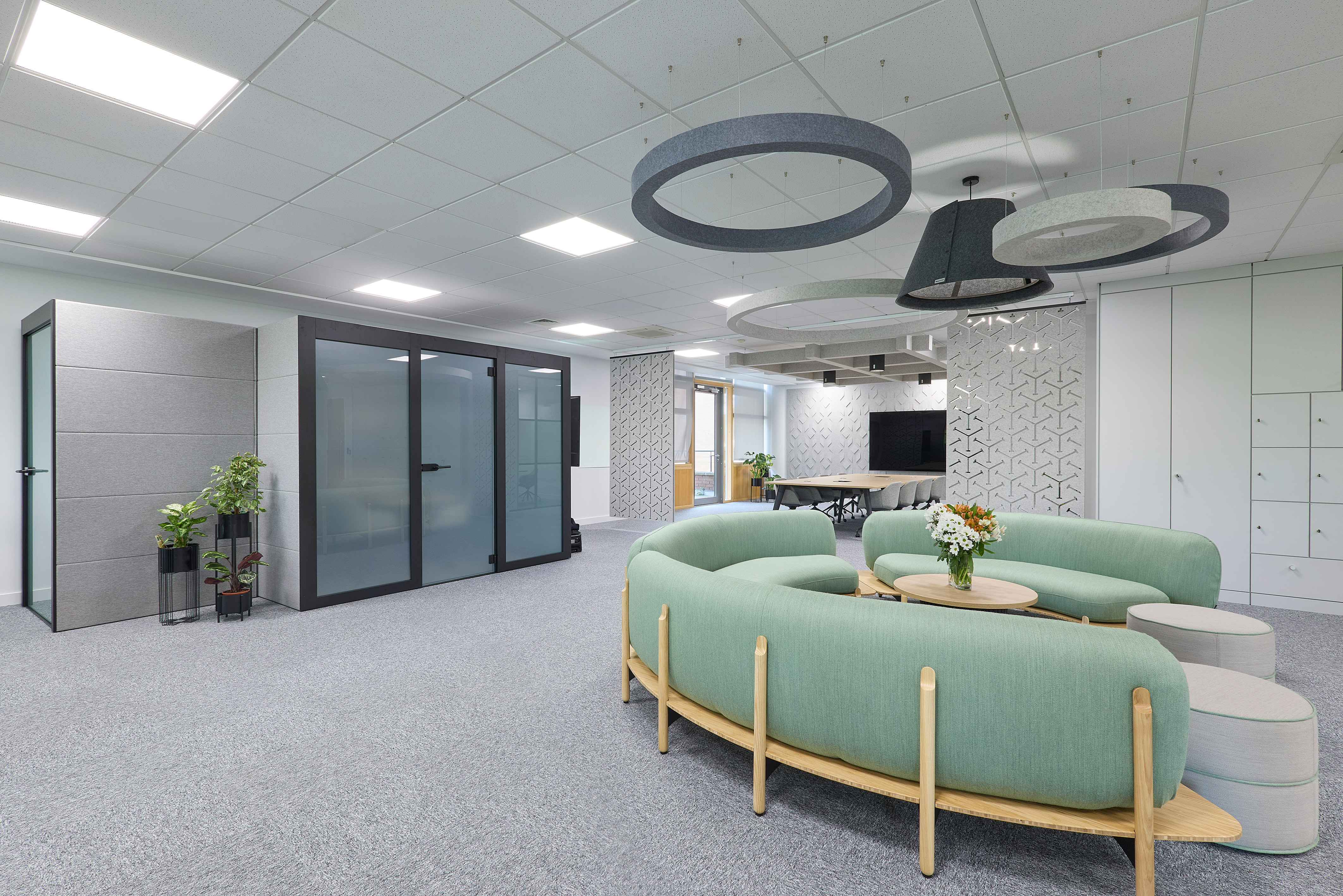 Modern office lounge features circular green sofas, coffee tables with flowers, and overhead ring lighting. Glass-walled meeting rooms and abstract wall designs create an open, stylish environment.