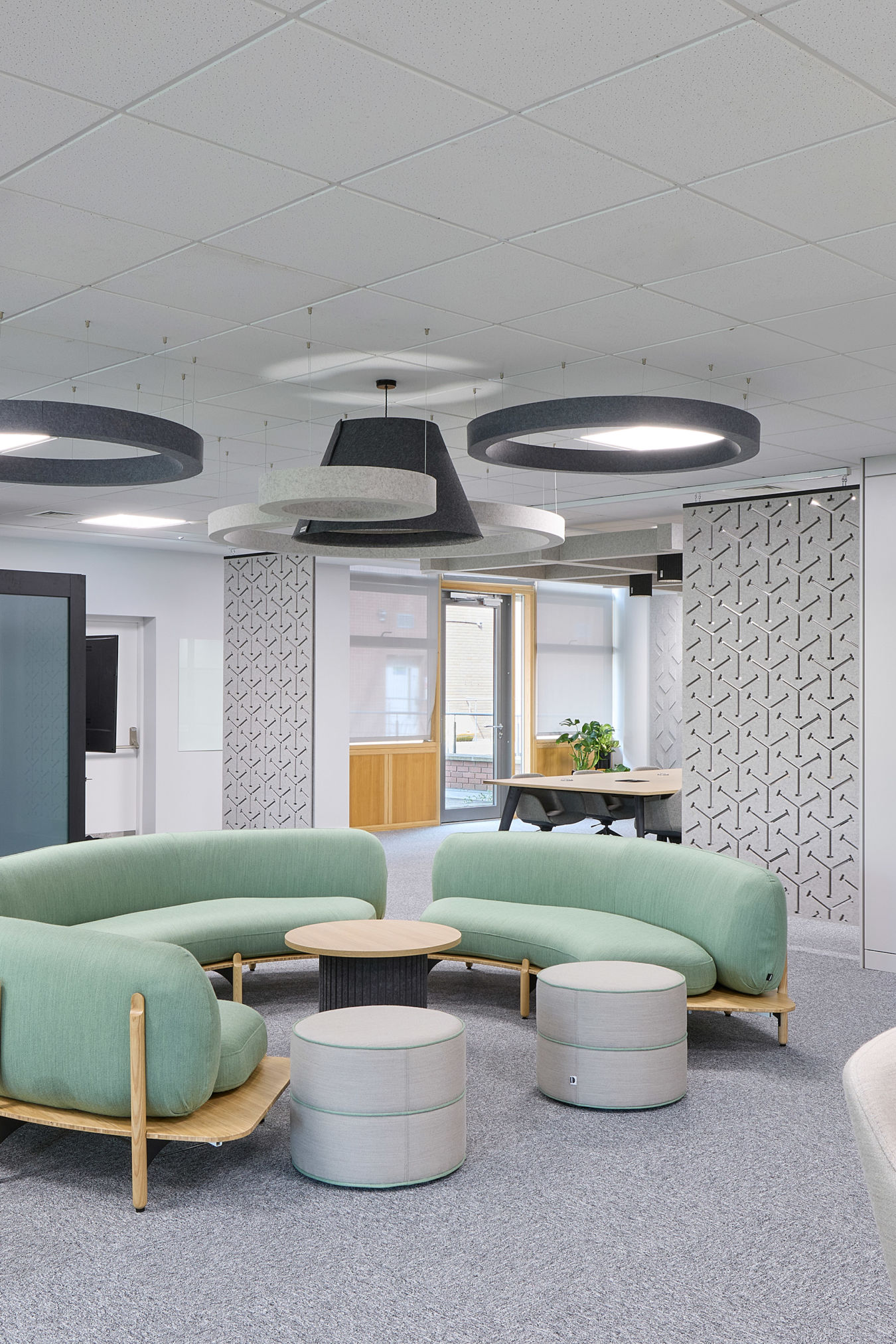 Modern office space featuring a curved green sofa set with round tables in the center, surrounded by chairs and lockers, under geometric ceiling lights on a gray carpeted floor.