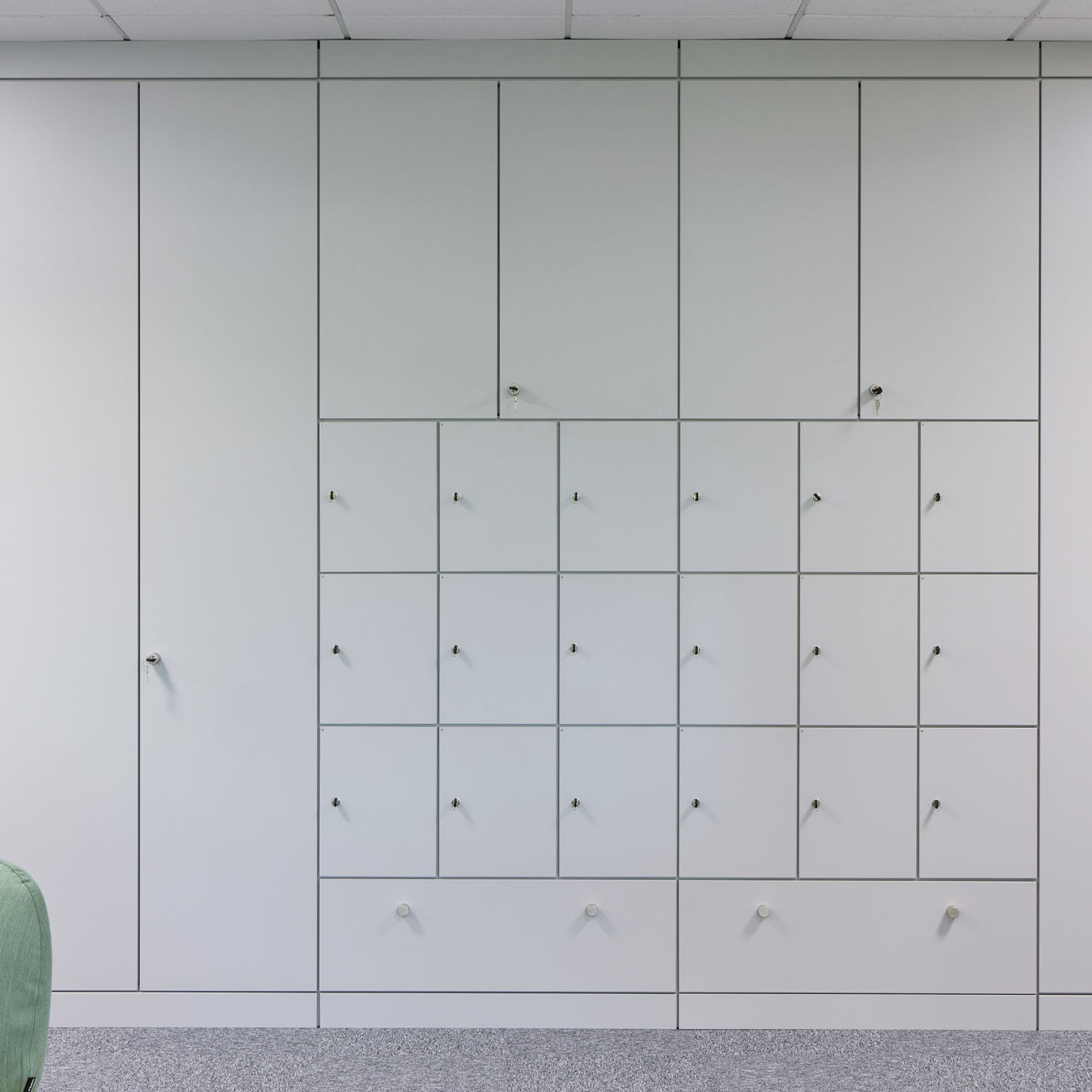 Modern office space with sleek white lockers and cabinets on a gray carpet. A light green couch and wooden slats add a touch of warmth