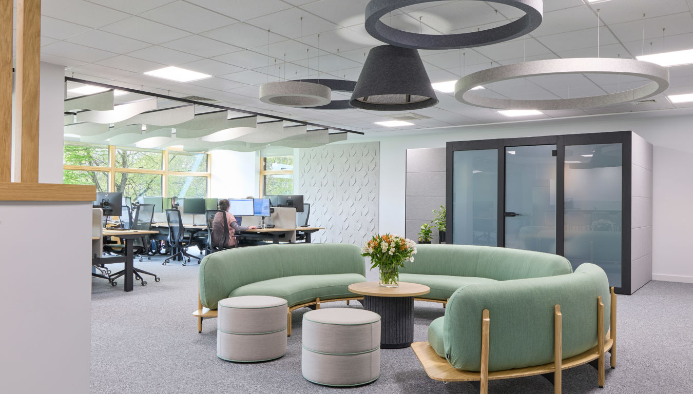 Modern office space with green circular sofa, round table, and stools in the foreground. Open-plan desks and greenery are seen in the background.