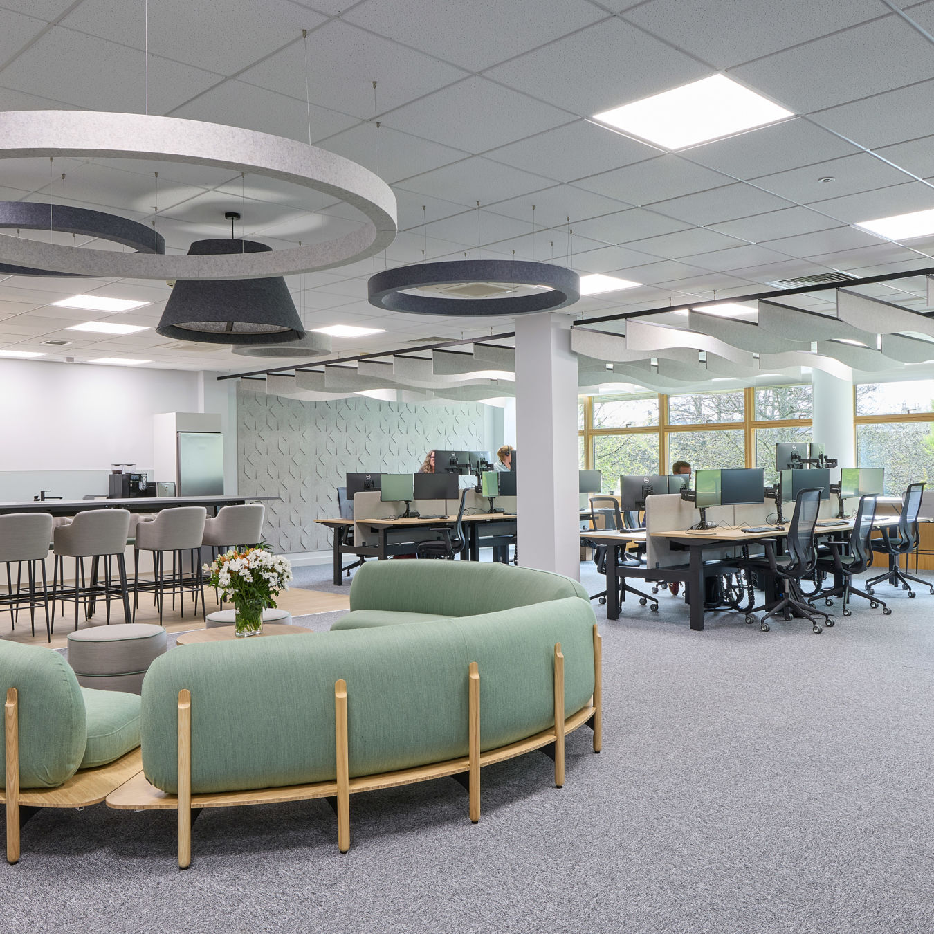 Modern office space with open layout, featuring green circular seating, pendant lights, workstations by large windows, and a person working at a desk.