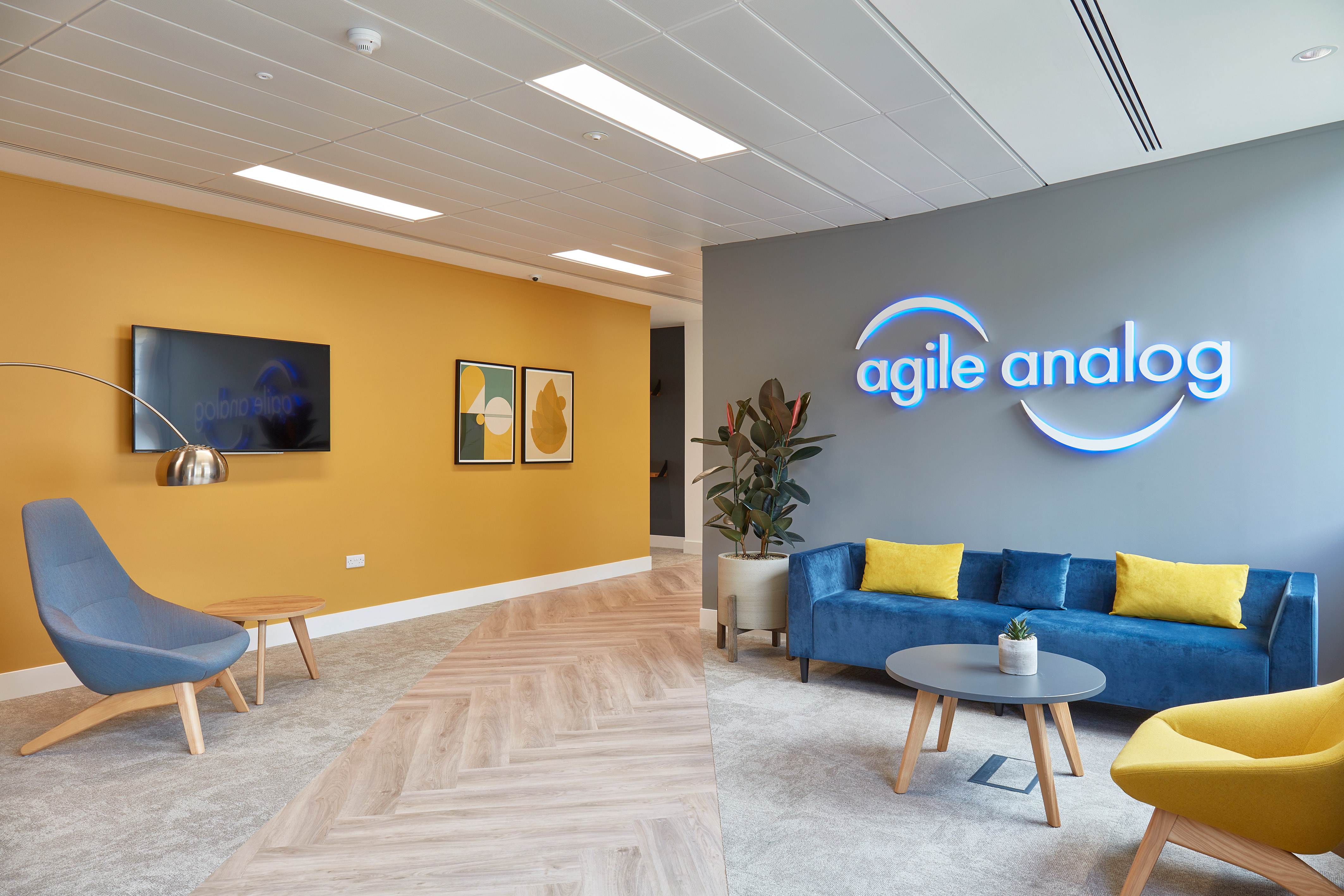 Lobby with a blue couch and yellow pillows under a neon "agile analog" sign, featuring a mustard accent wall with framed art. A blue chair and wooden table complete the setting.