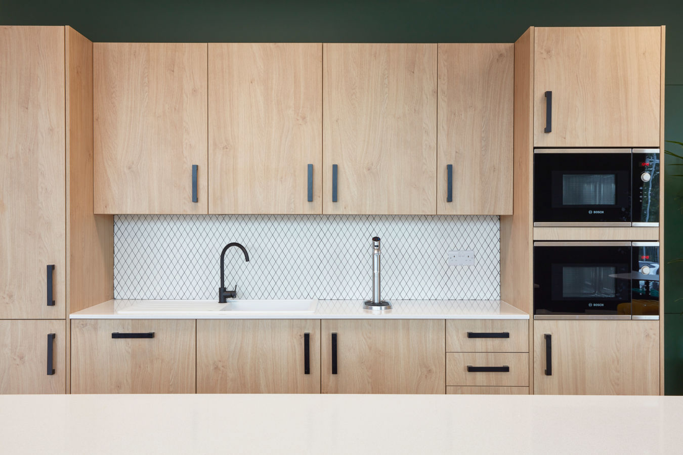 Wooden kitchen cabinets with black handles surround a white countertop featuring a black faucet. The backsplash has a diamond pattern. Two Bosch ovens are built into the right side.