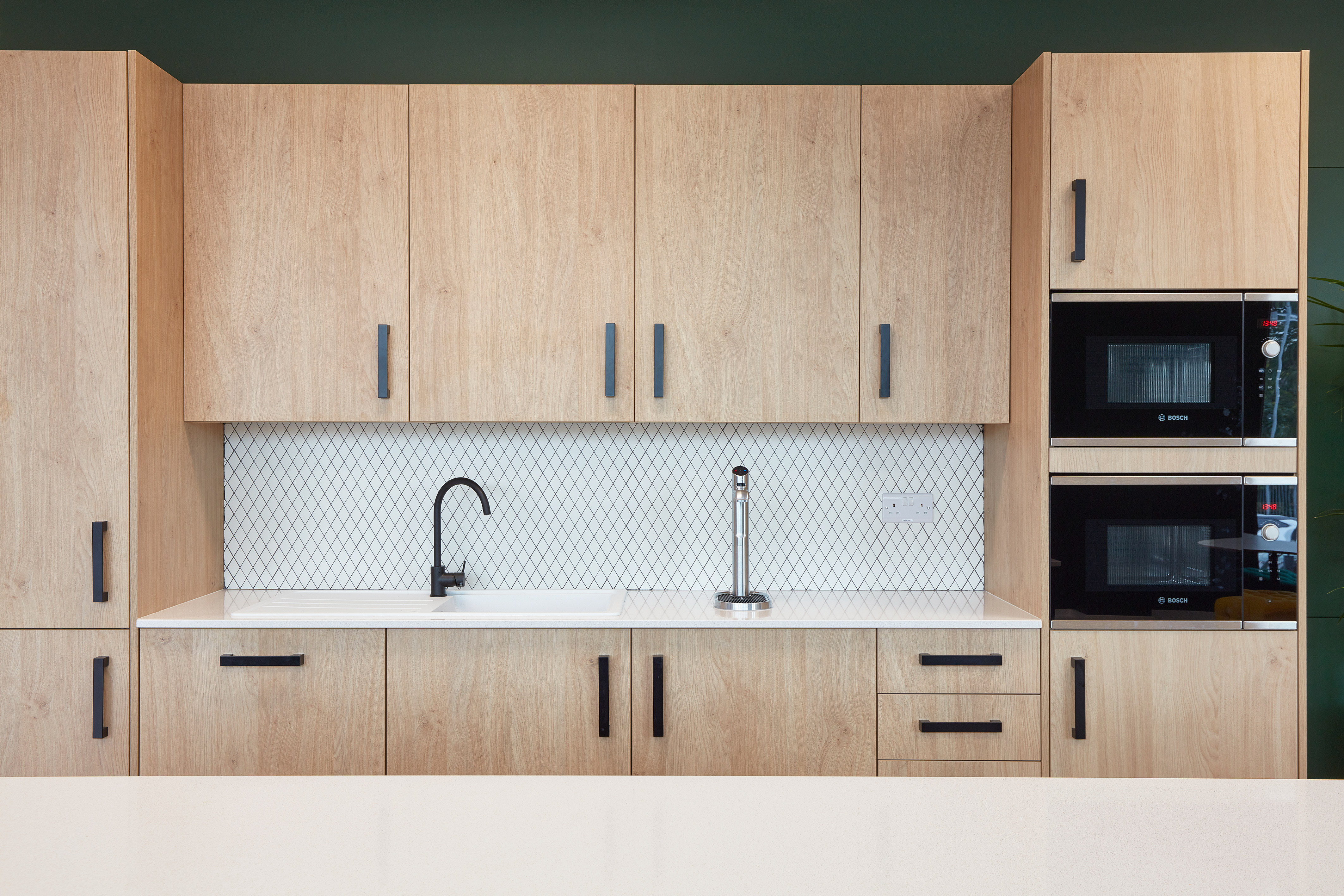 Wooden kitchen cabinets with black handles surround a white countertop featuring a black faucet. The backsplash has a diamond pattern. Two Bosch ovens are built into the right side.