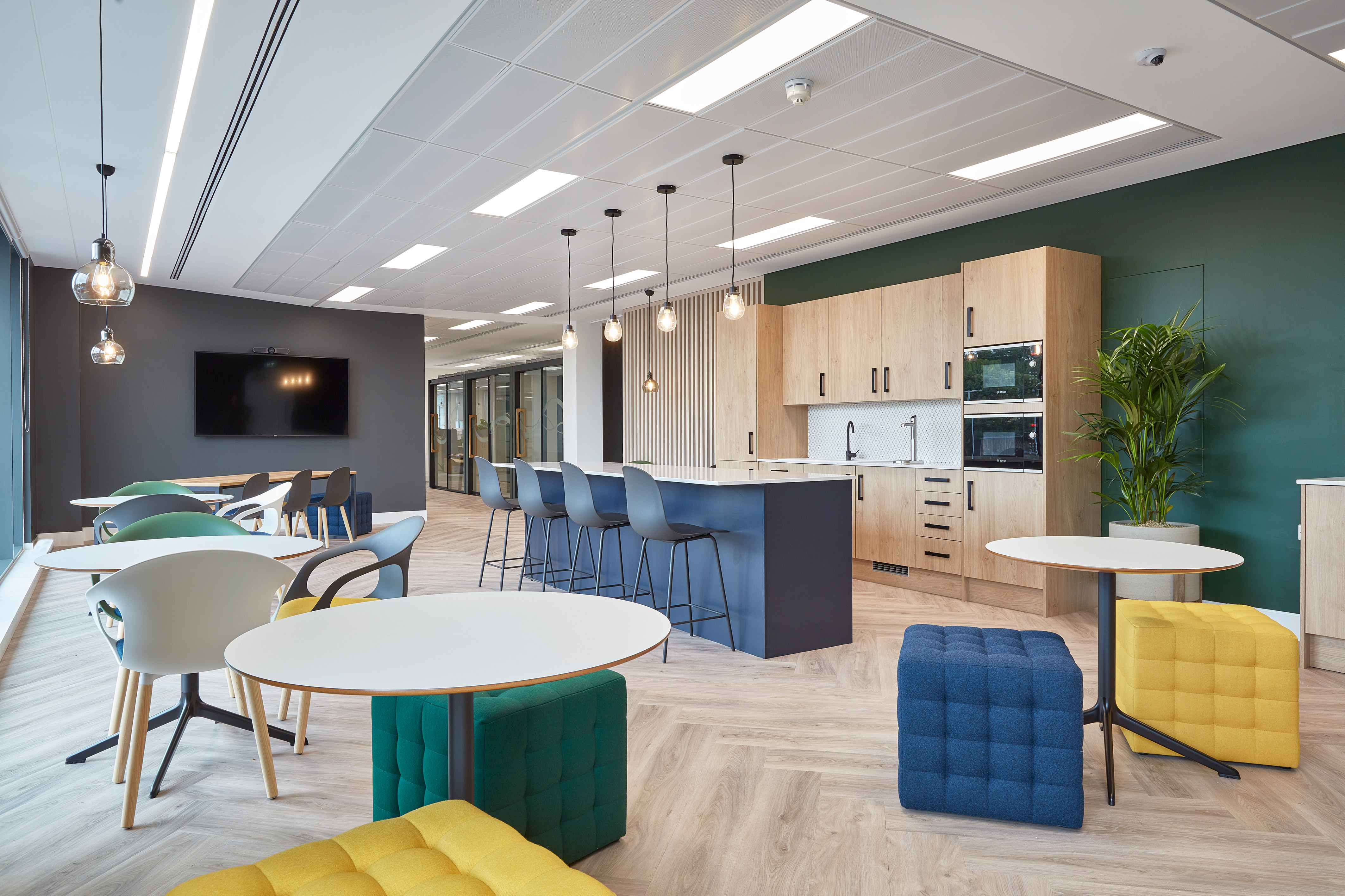 Modern office break room with round tables, colorful chairs, and blue barstools at a central island. Wooden cabinets and appliances line one wall, with a flat-screen TV and hanging lights.