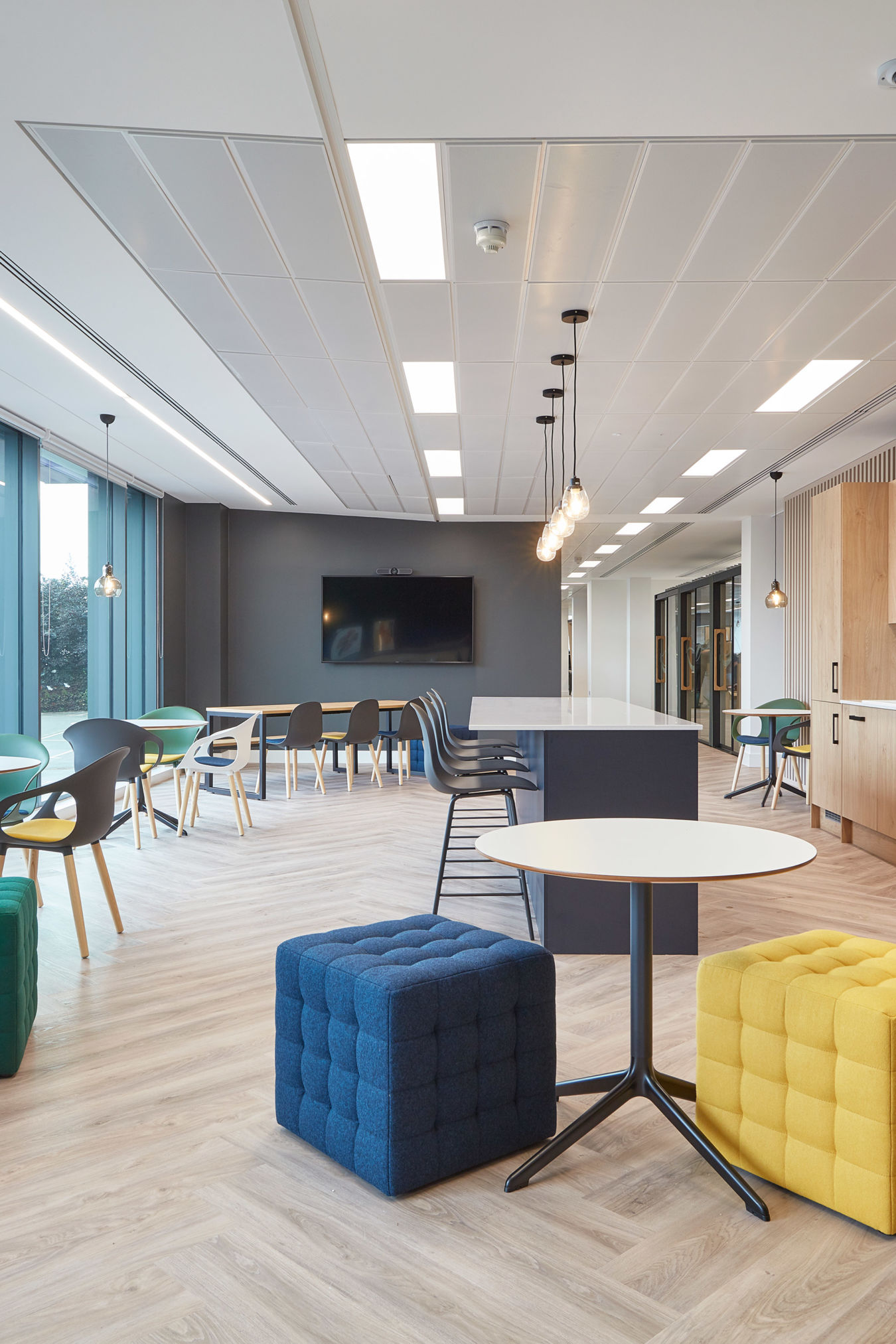 Modern office lounge with colorful seating, round tables, and pendant lights; a flat-screen TV is mounted on a dark wall, surrounded by windows and natural light.