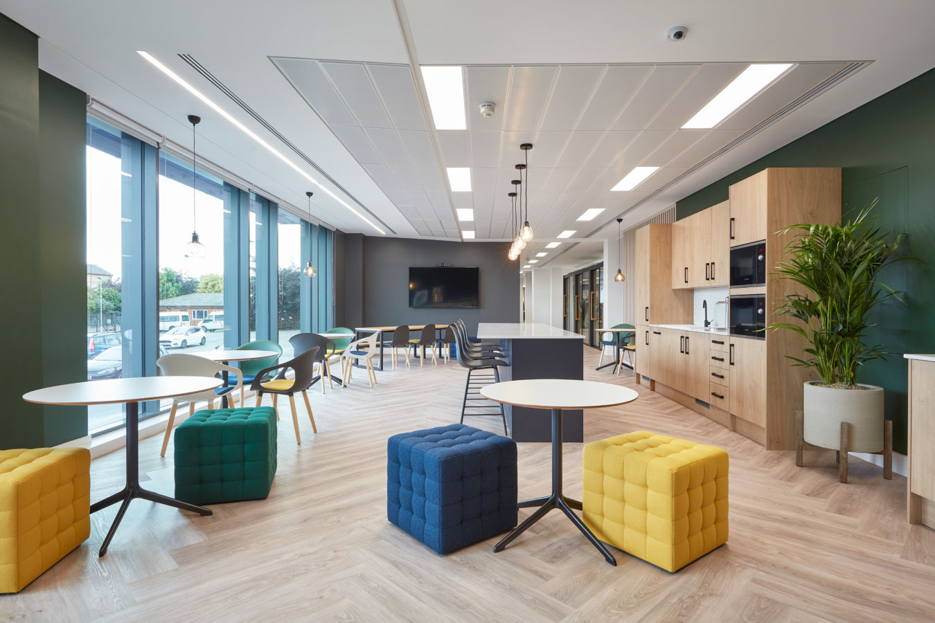 Modern office lounge with colorful seating, round tables, and pendant lights; a flat-screen TV is mounted on a dark wall, surrounded by windows and natural light.