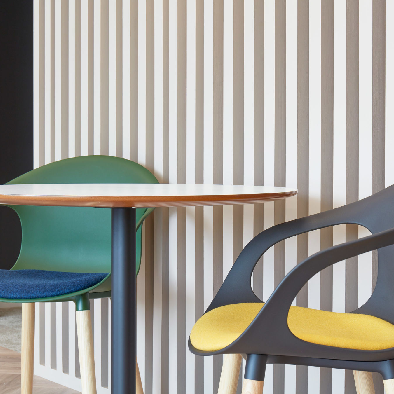 Two modern chairs with colorful cushions sit beside a small round table against a wall featuring vertical beige and white stripes.