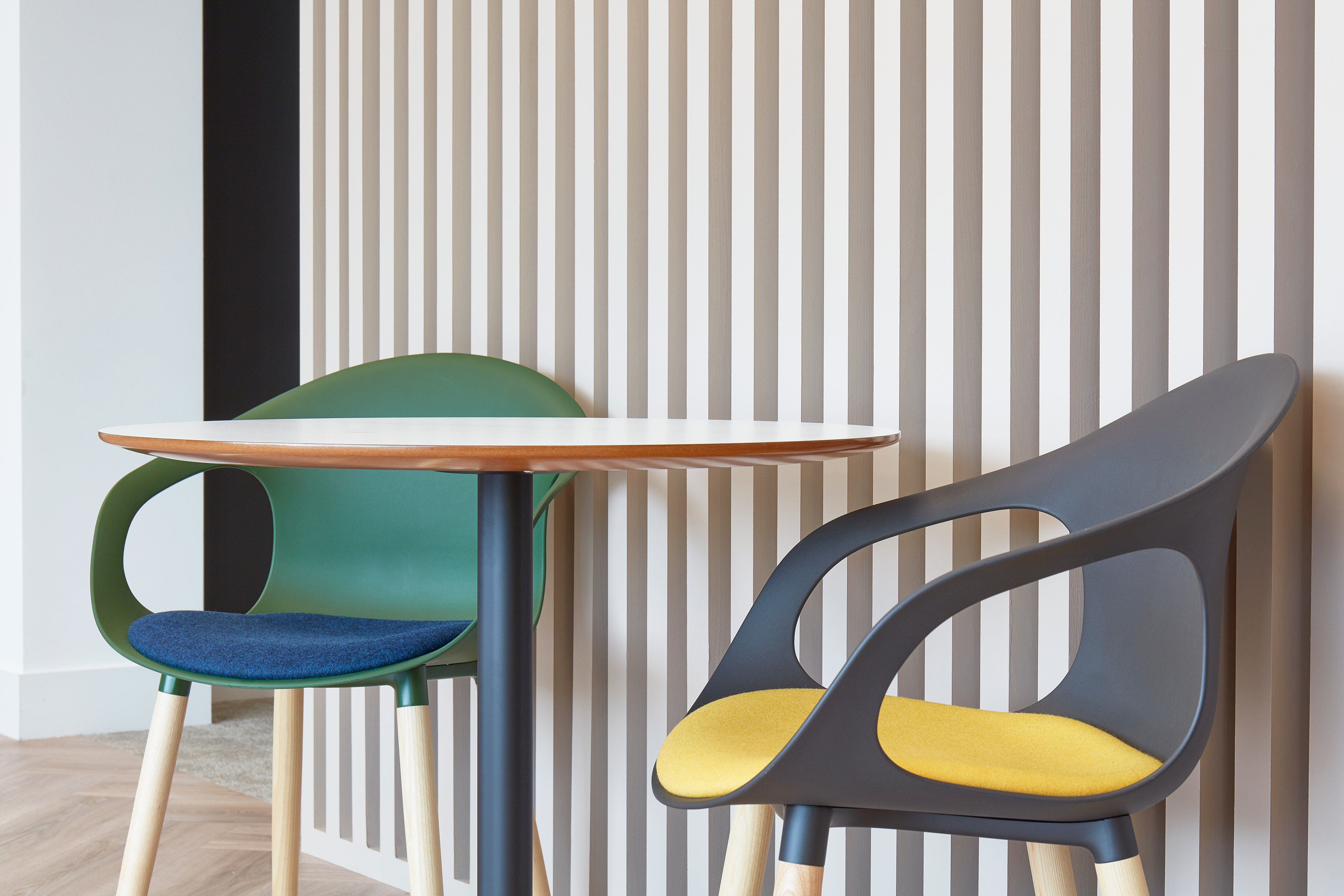 Two modern chairs with colorful cushions sit beside a small round table against a wall featuring vertical beige and white stripes.