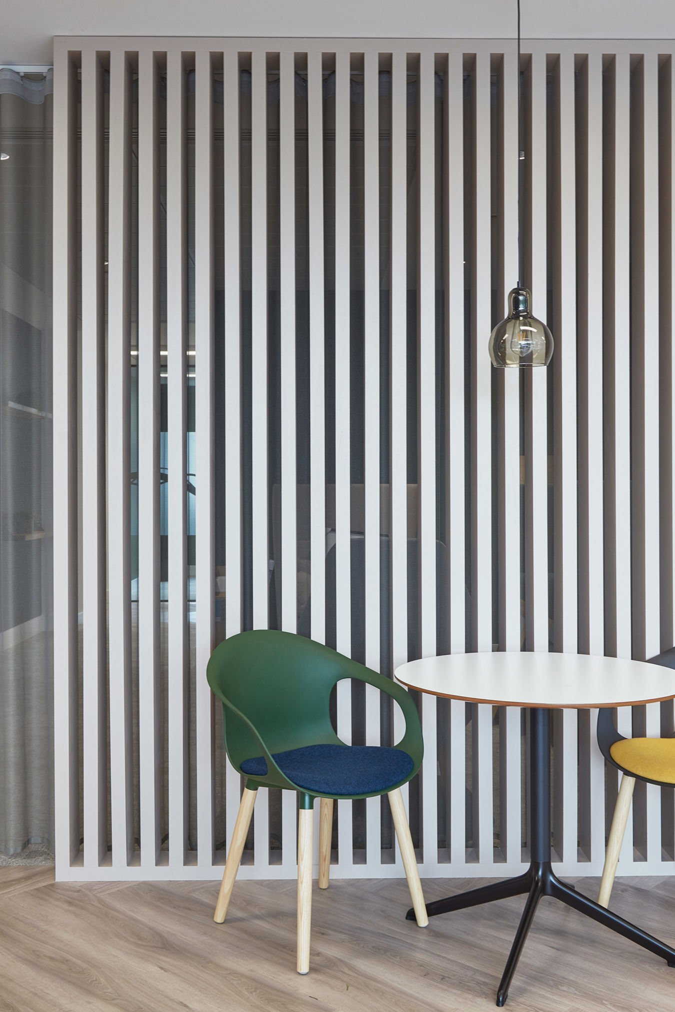 Two colorful chairs face a small round table under a single pendant light, set against a vertical slat partition in a modern, minimalist room.