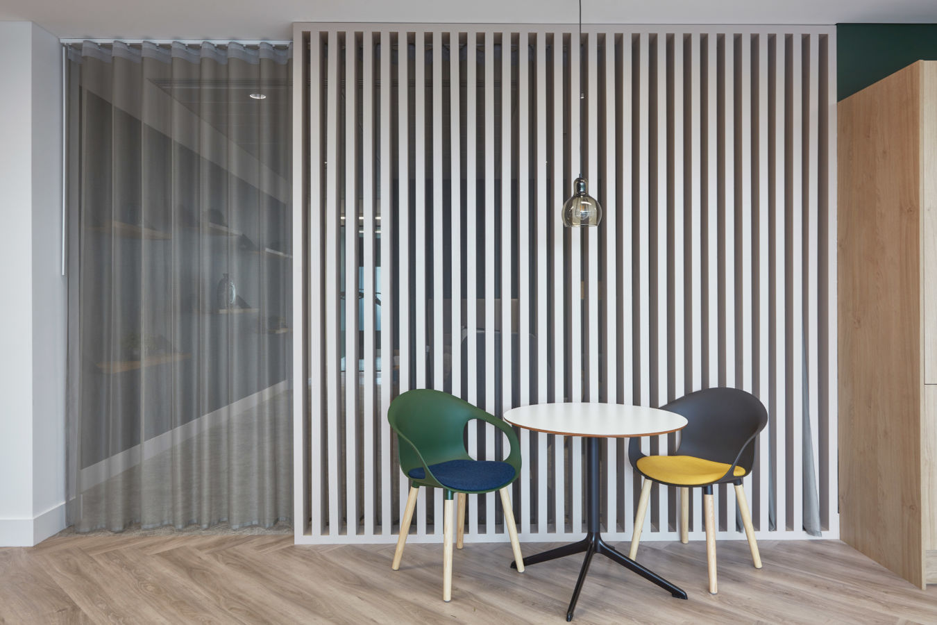 Two colorful chairs face a small round table under a single pendant light, set against a vertical slat partition in a modern, minimalist room.