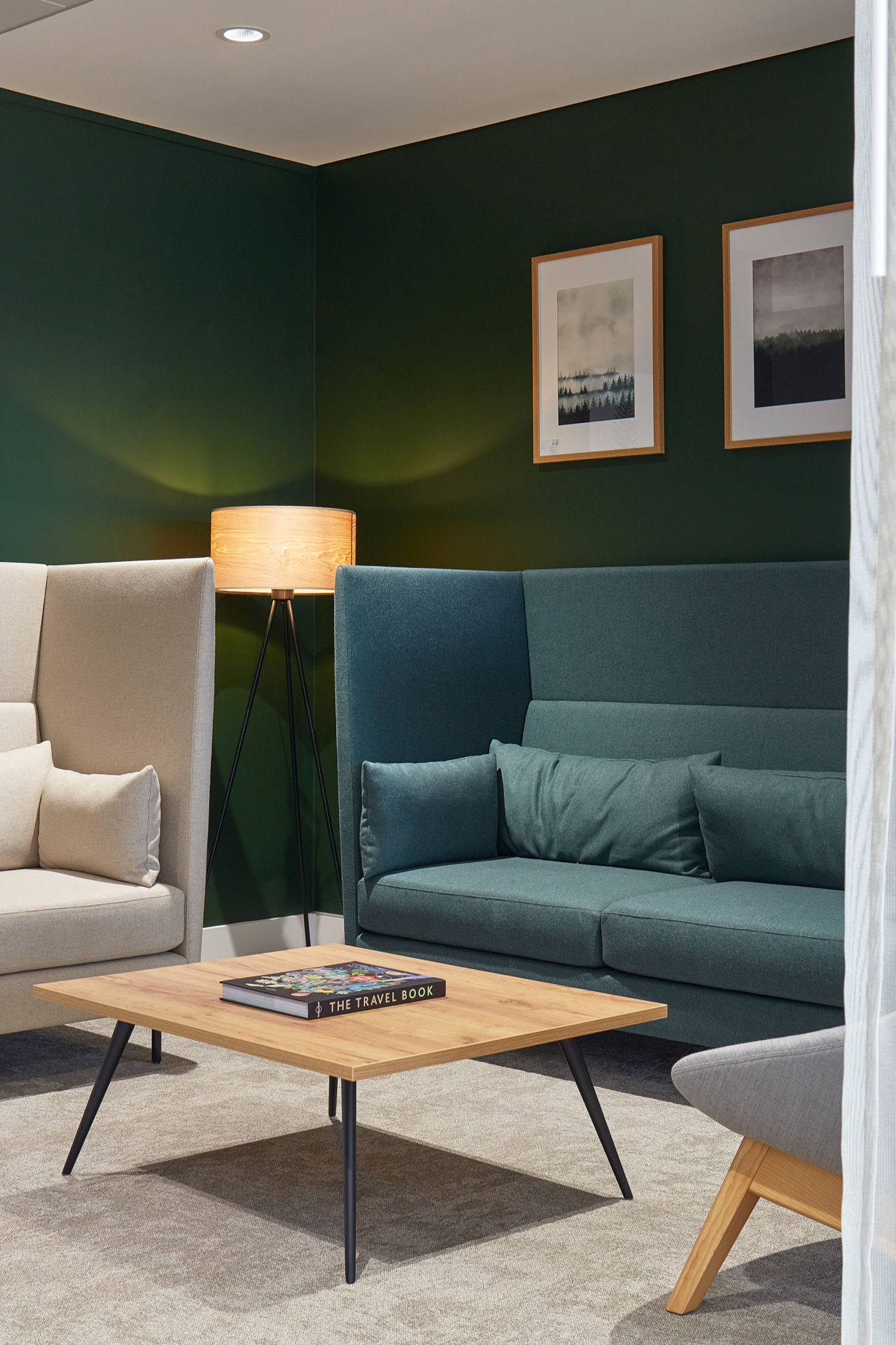 Two sofas face each other beside a wooden coffee table with "THE TRAVEL BOOK" in a cozy room; a tripod lamp is lit, and paintings hang on dark green walls.