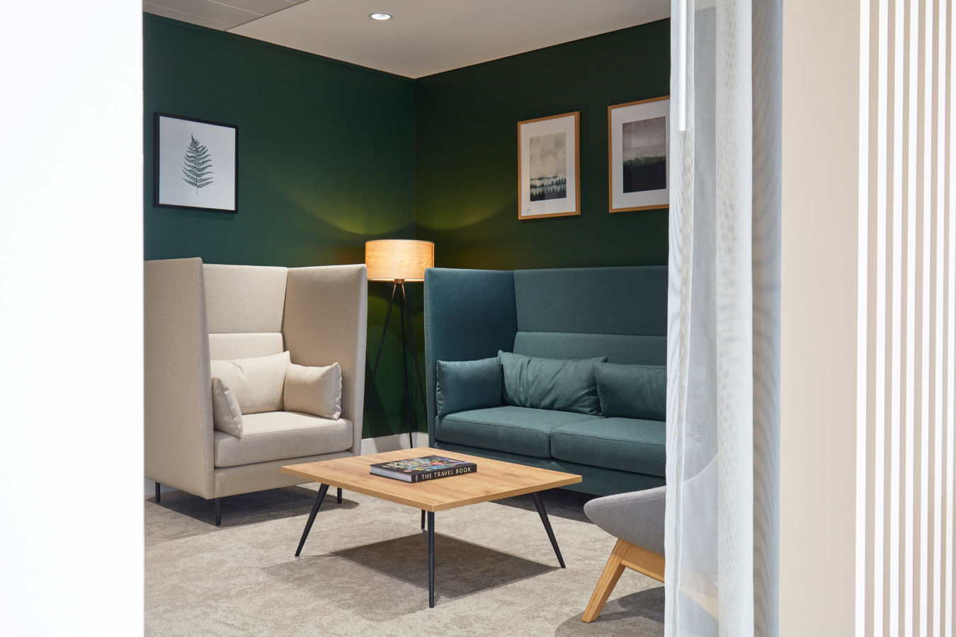 Two sofas face each other beside a wooden coffee table with "THE TRAVEL BOOK" in a cozy room; a tripod lamp is lit, and paintings hang on dark green walls.