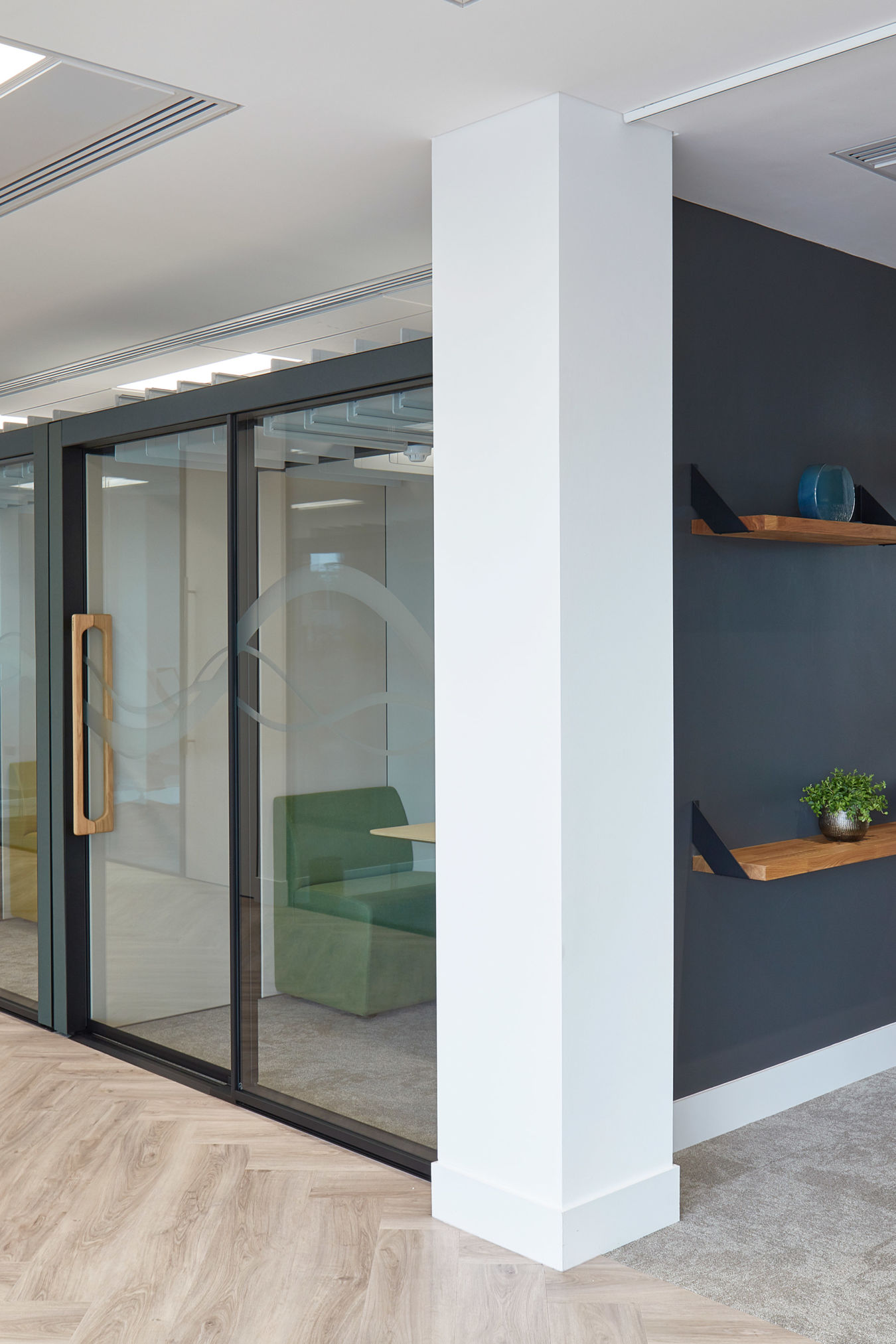 Glass office door with wooden handle, revealing green seating area inside, stands near dark grey wall with floating shelves and decorative items, set in modern interior space.