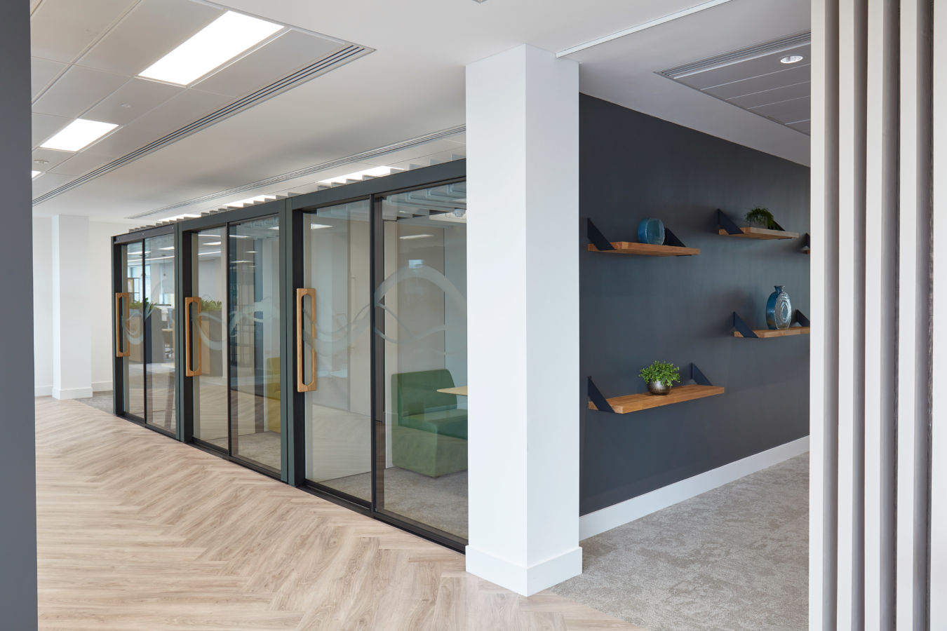 Glass office door with wooden handle, revealing green seating area inside, stands near dark grey wall with floating shelves and decorative items, set in modern interior space.