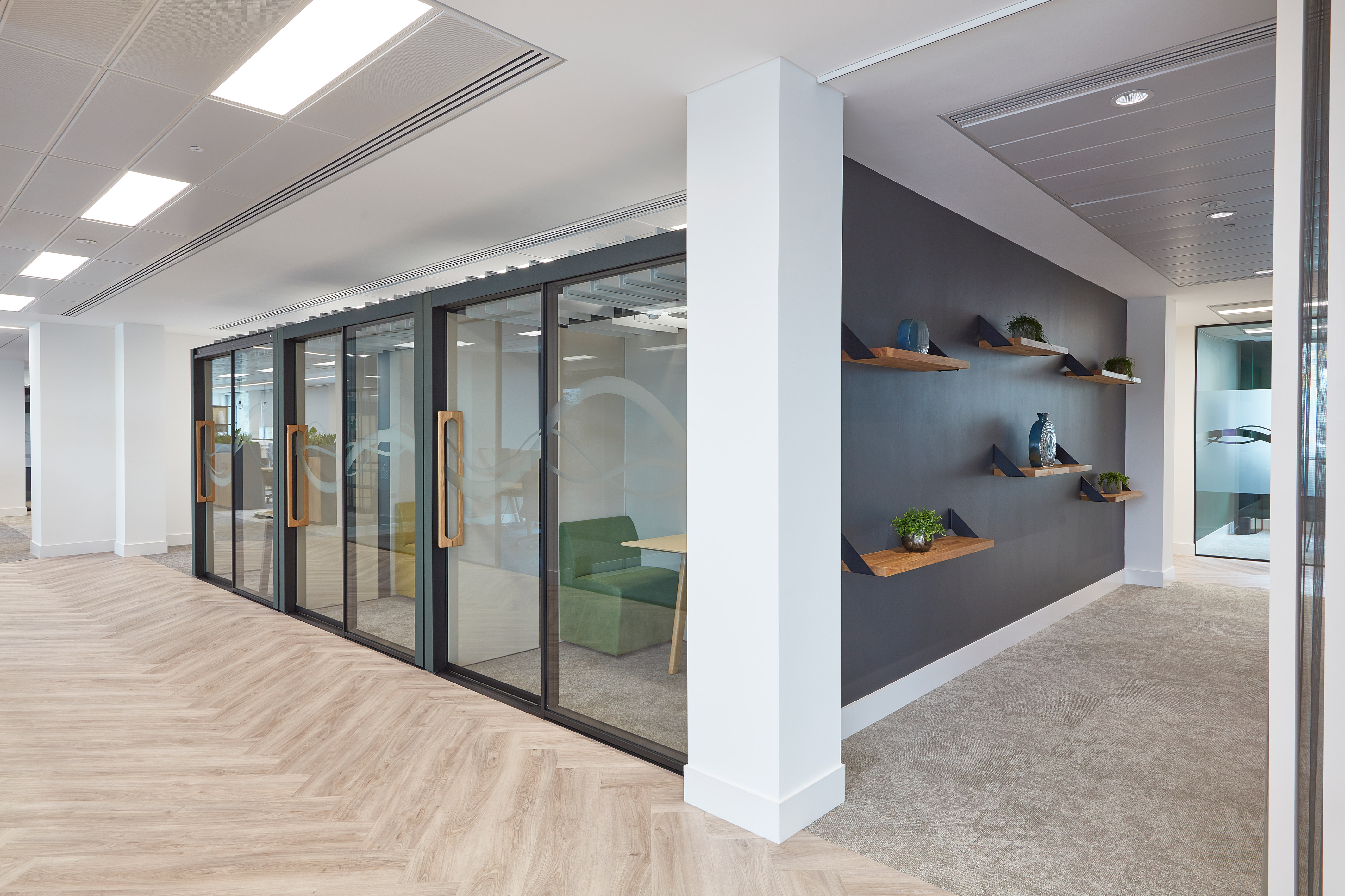 Glass-walled meeting rooms with wooden handles line a modern office hallway; opposite, a dark wall with wooden shelves holds decorative vases and plants, featuring light wood and carpet flooring.