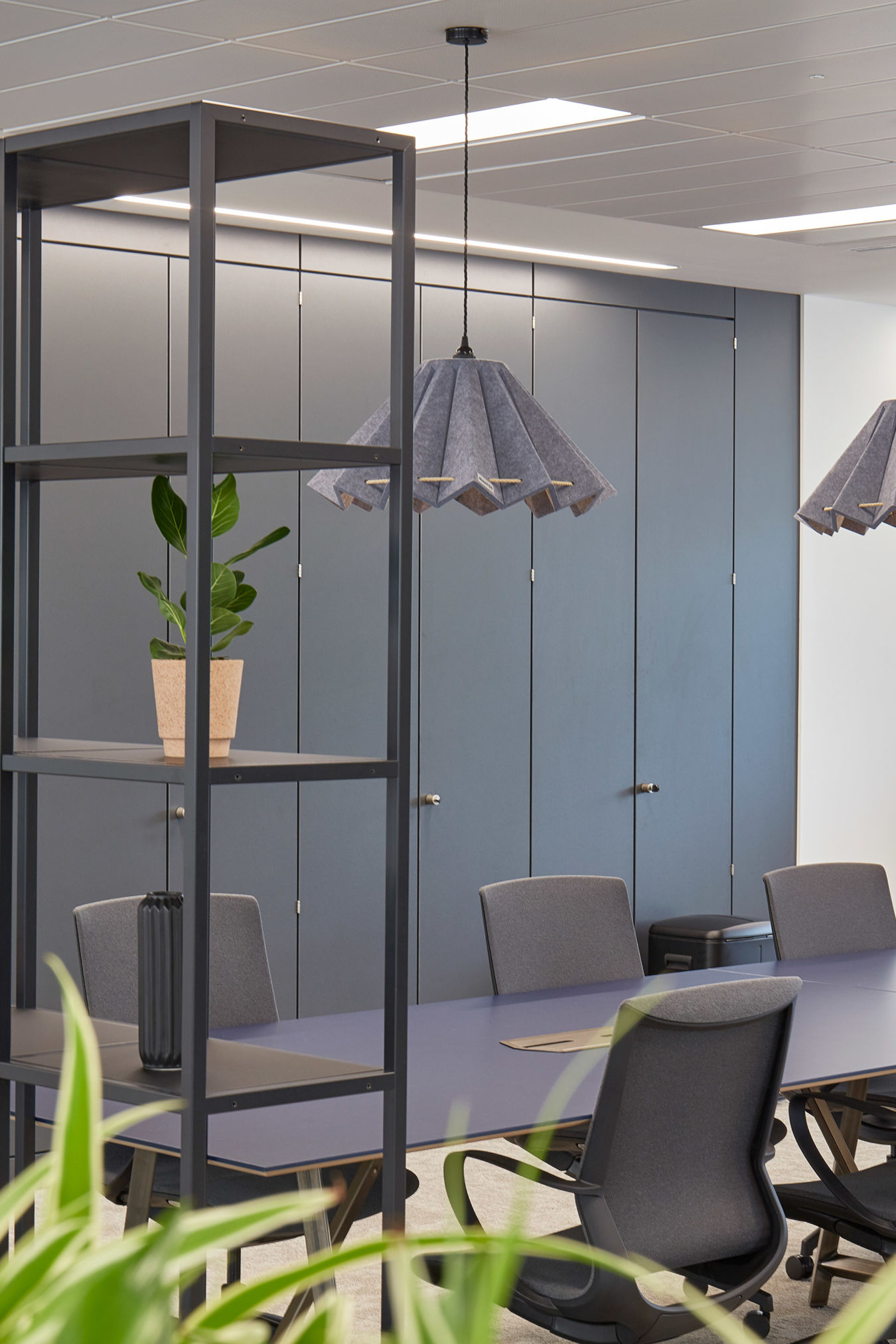 A modern office meeting room with a long table, surrounded by ergonomic chairs, overhead pendant lights, and open shelving with decorative plants, featuring gray cabinets in the background.