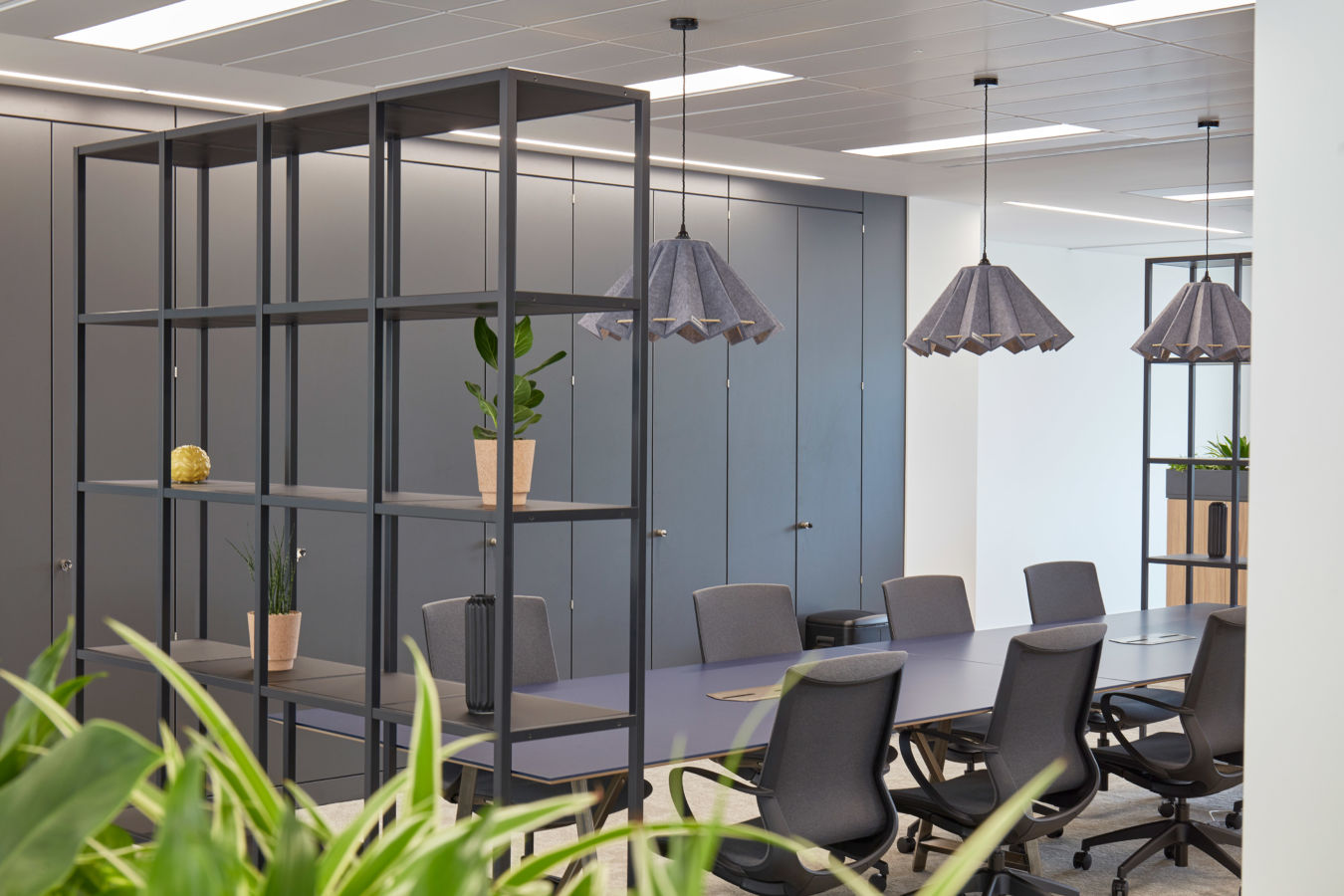 A modern office meeting room with a long table, surrounded by ergonomic chairs, overhead pendant lights, and open shelving with decorative plants, featuring gray cabinets in the background.
