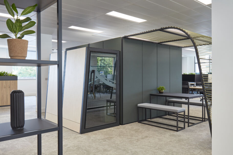 A modern office meeting pod with glass walls and a bench seating area is surrounded by an open-plan workspace with plants and minimalistic decor.