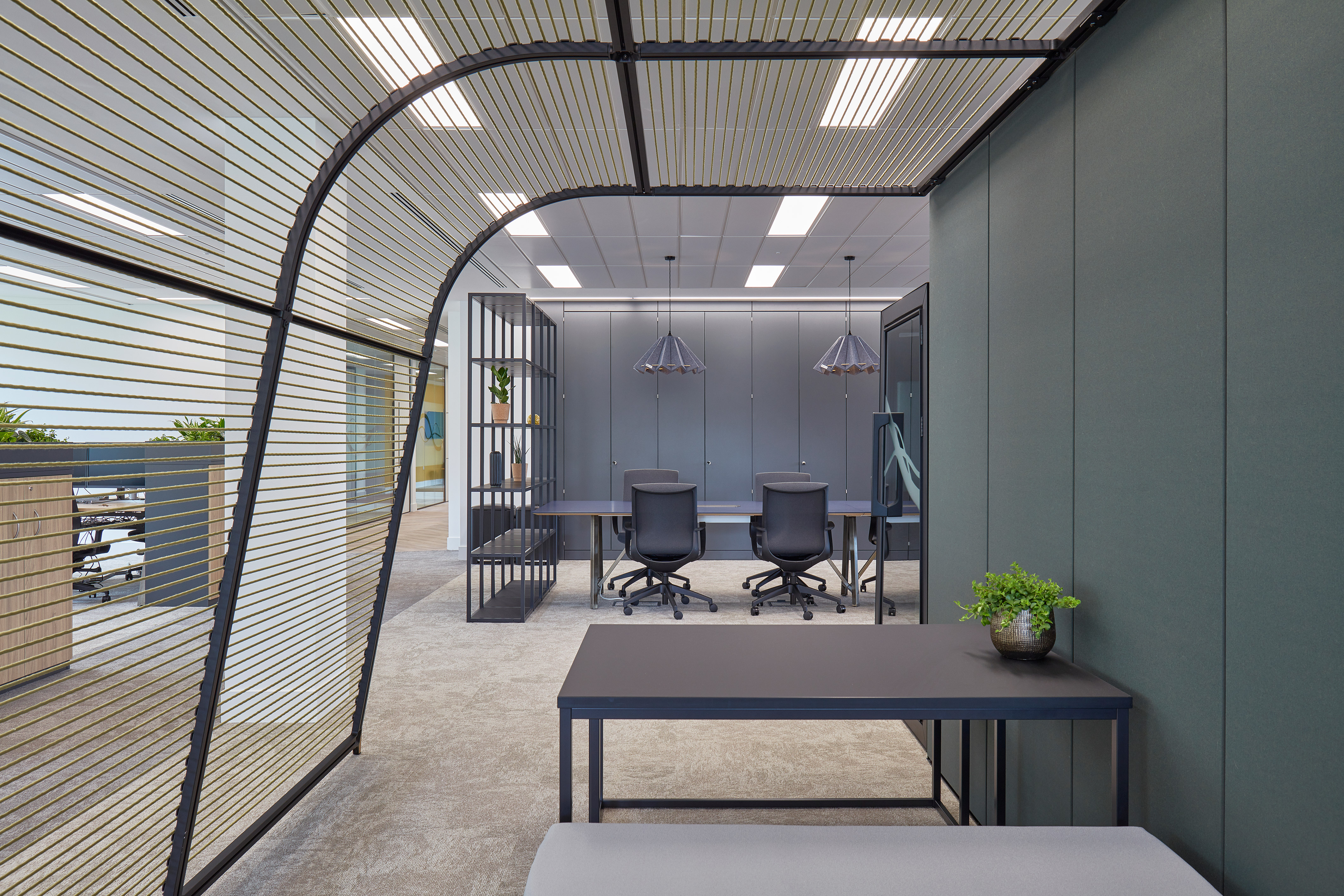 Modern office space with two black chairs and a table facing grey cabinets; features overhead lighting, a potted plant on a side table, and a striped partition wall.
