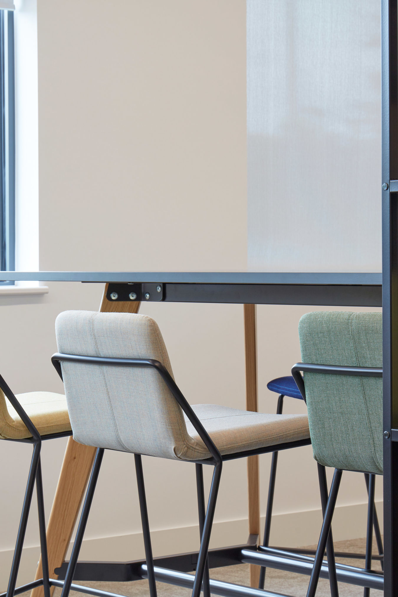 Four high-back chairs in pastel colors are neatly arranged around a sleek black table in a modern, minimalist room with white walls and a small window.