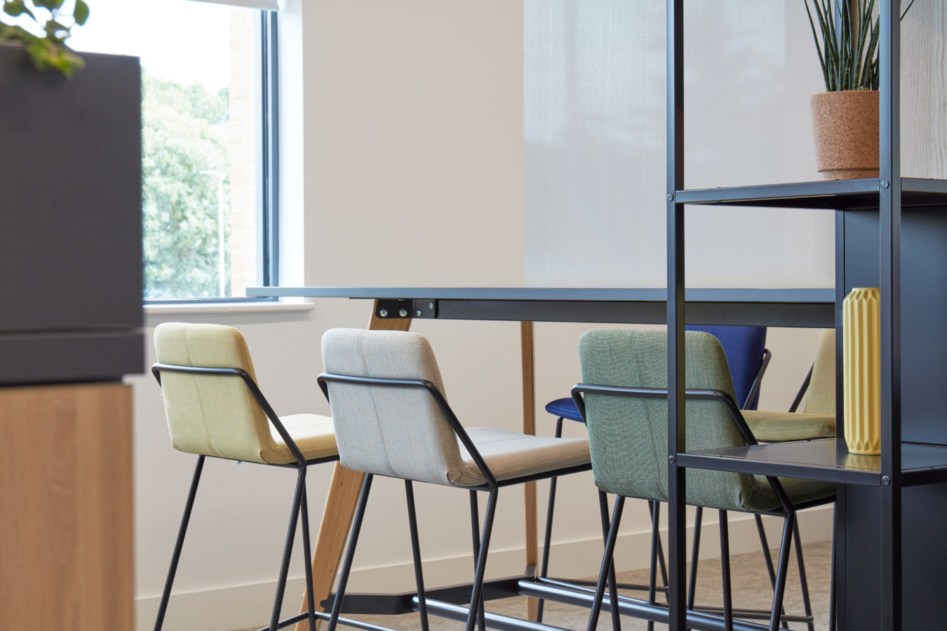 Four high-back chairs in pastel colors are neatly arranged around a sleek black table in a modern, minimalist room with white walls and a small window.