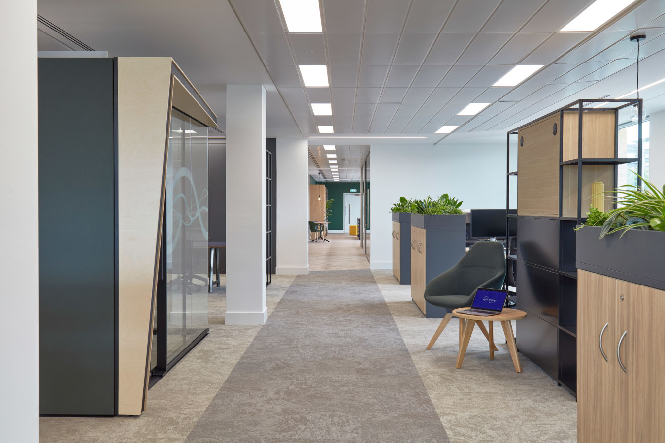 Modern office hallway with work pods and plant-topped cabinets on either side; a chair and small table with an open laptop are on the right, under bright ceiling lights.