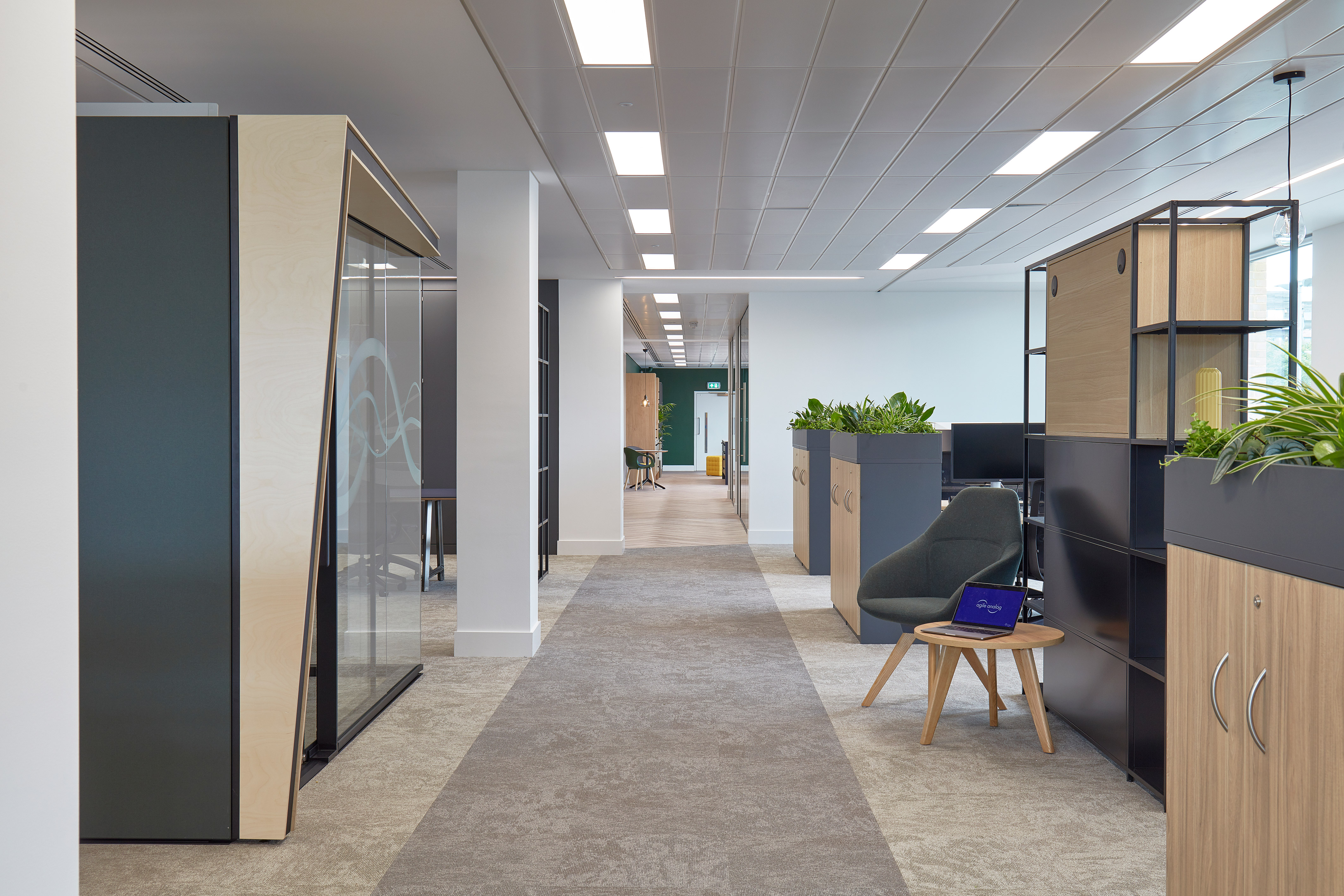 Modern office hallway with work pods and plant-topped cabinets on either side; a chair and small table with an open laptop are on the right, under bright ceiling lights.