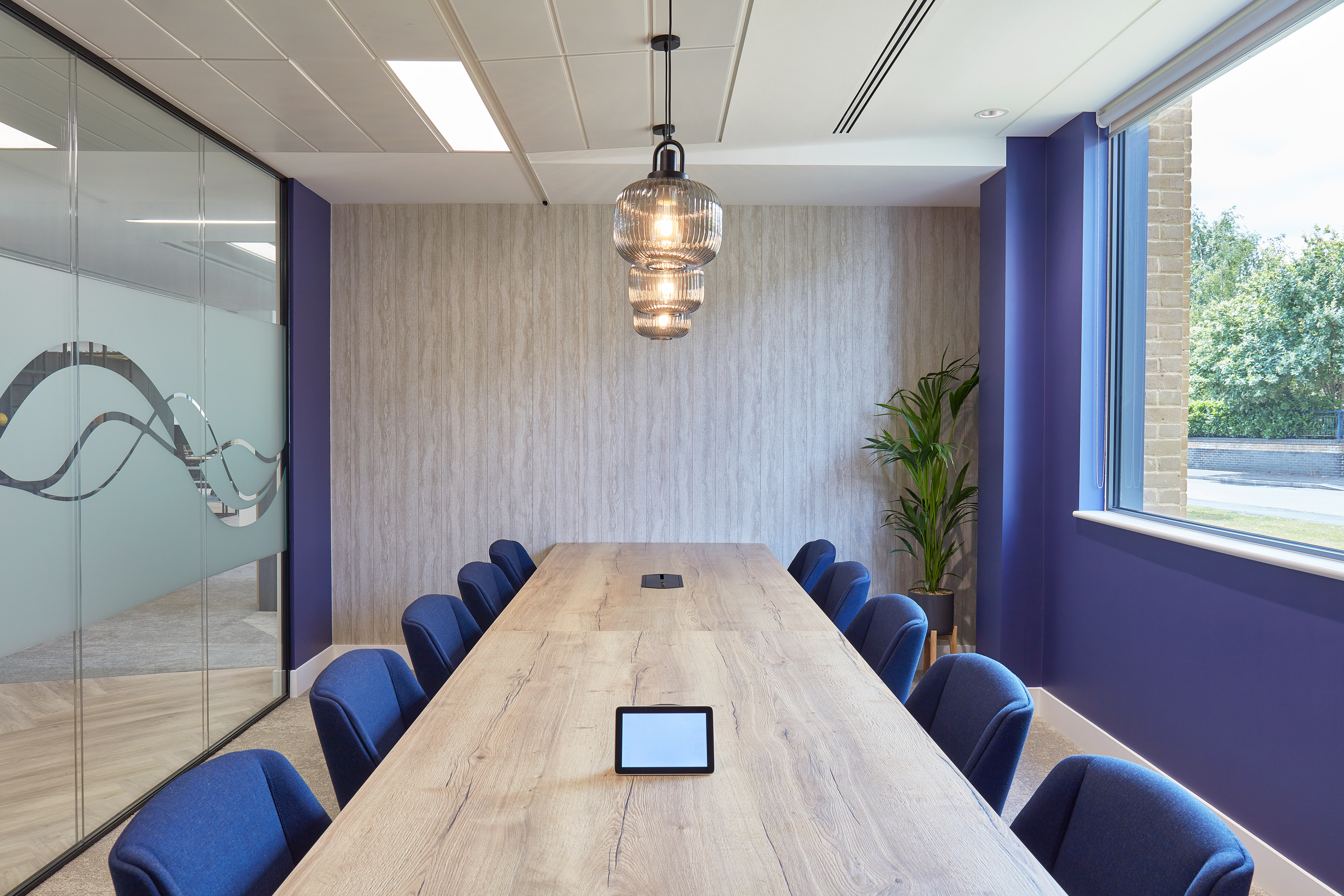 Conference table with blue chairs and glass lamps overhead, situated in a modern meeting room with glass walls and a window view of greenery.