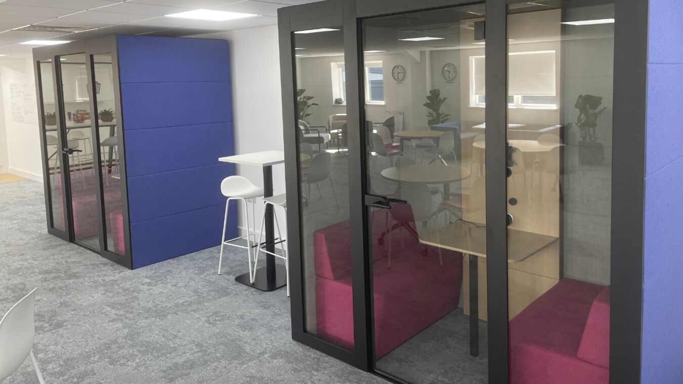 Glass-walled office pods with purple exterior feature pink cushioned seating inside, situated in an open-plan office with a standing desk and white stool nearby.
