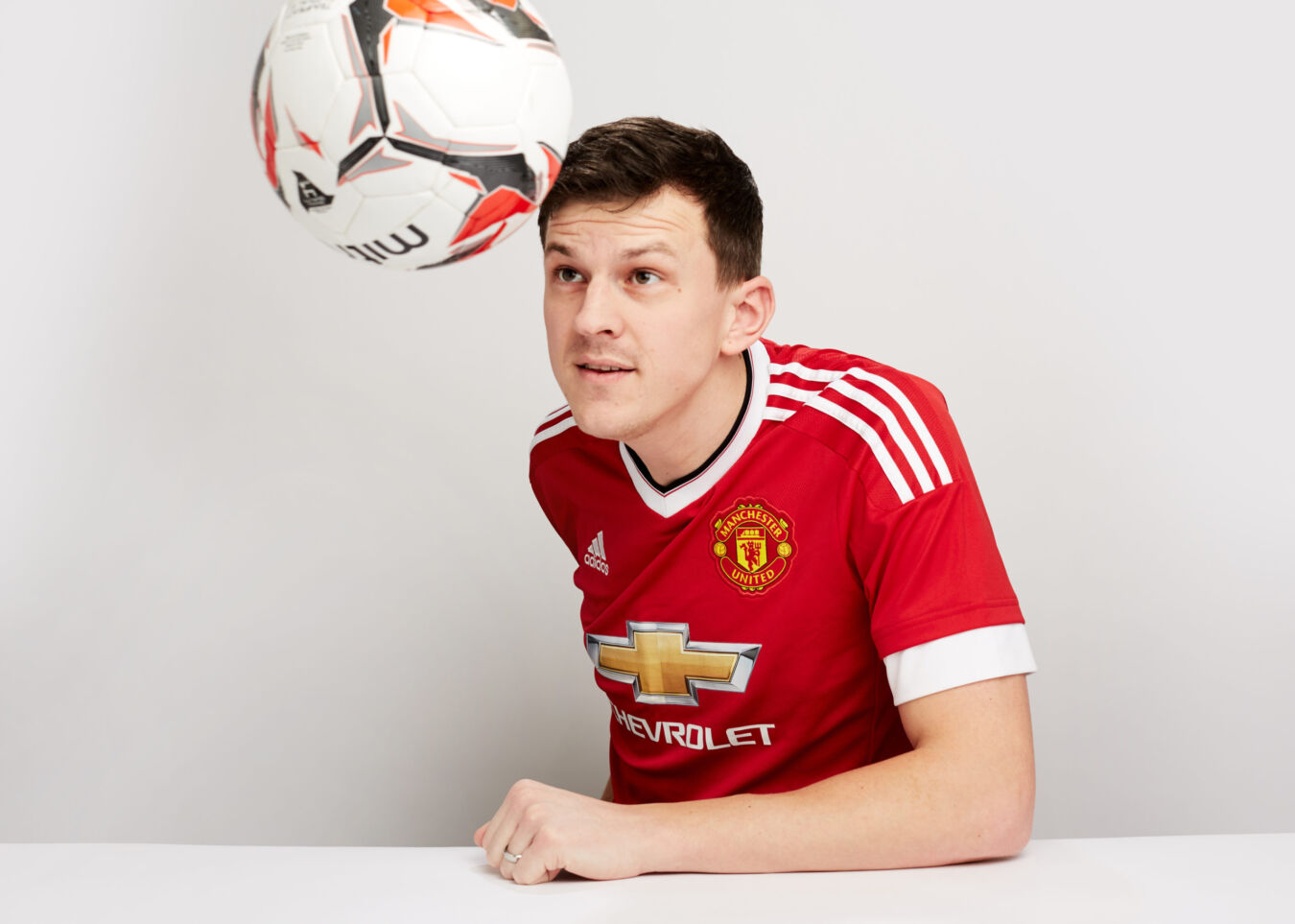A person in a red Manchester United jersey focuses intently on an airborne soccer ball against a plain white background, showing Adidas and Chevrolet logos.