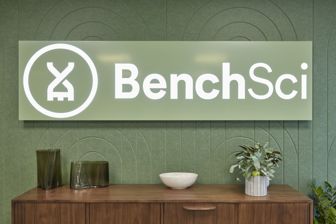 Sign with the text "BenchSci" and a circular logo, displayed on a green patterned wall. Below, a wooden cabinet holds decorative glass vases, a bowl, and a potted plant.