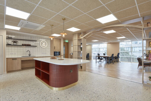 Modern office with an open-plan layout featuring a tiled kitchen area, central island with red accents, wooden shelves, and large glass windows