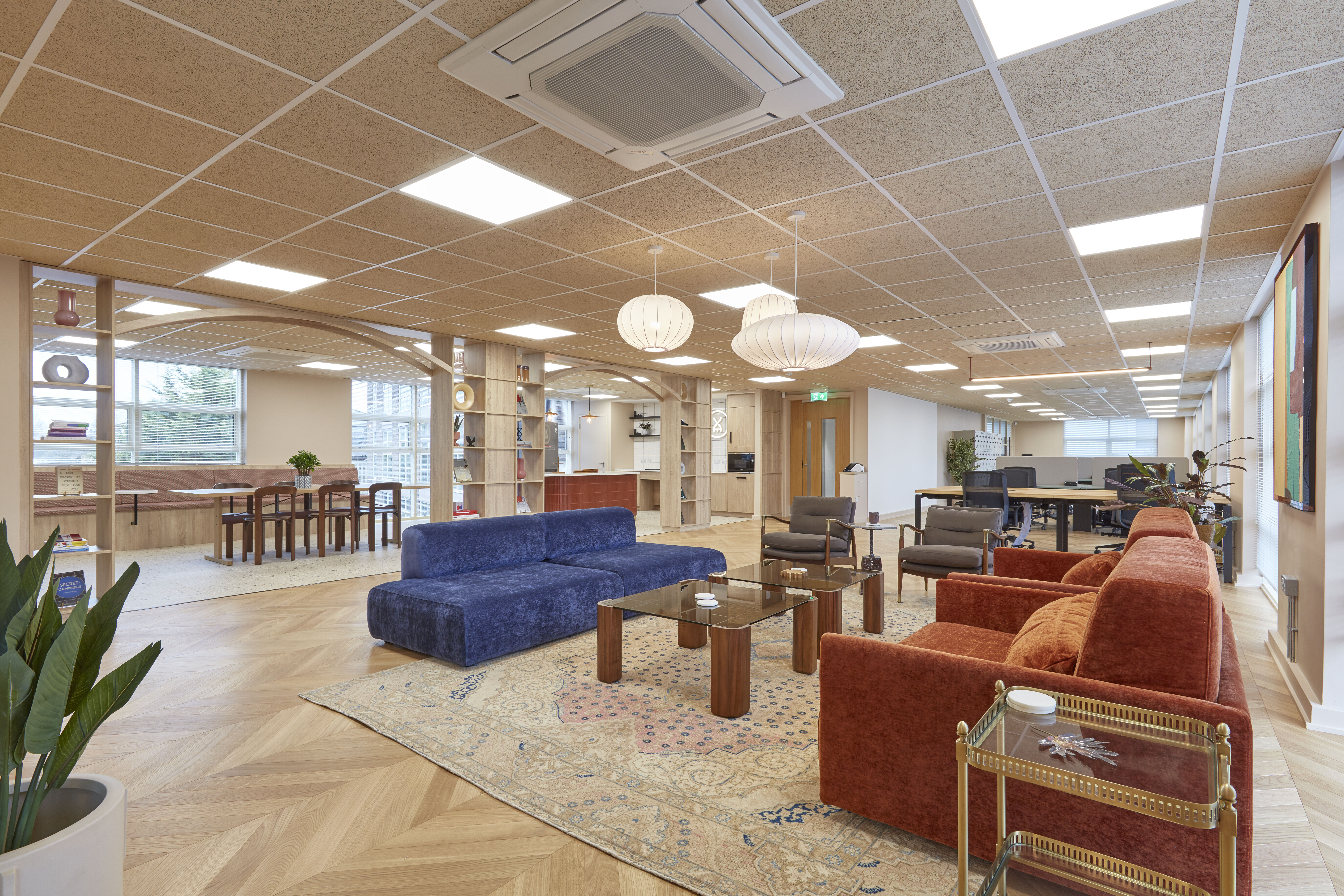 Spacious office with modern decor; features large blue and red sofas, wooden tables, pendant lights, and a patterned rug, creating a warm and inviting atmosphere