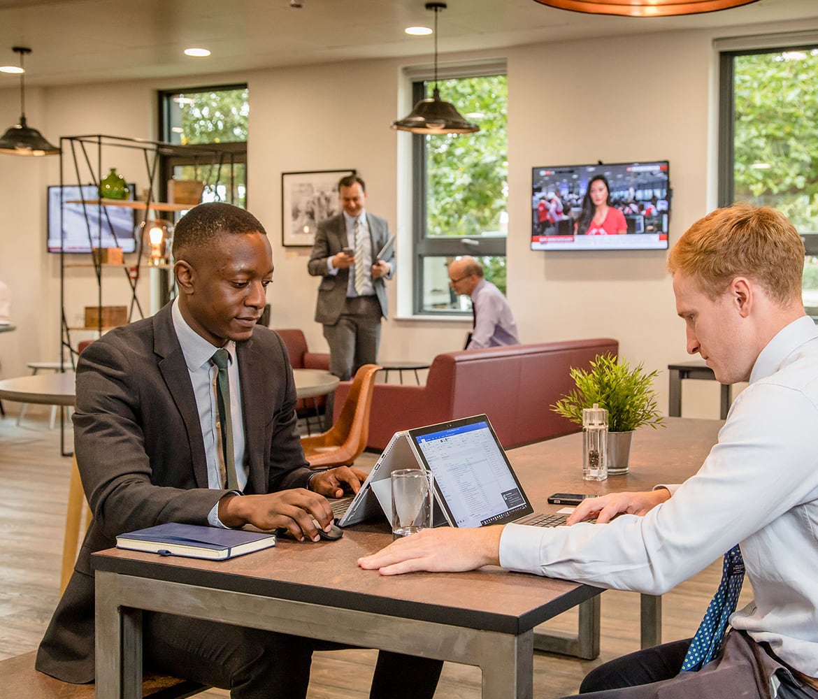 Two men in business attire work on laptops at a table in a modern office lounge, with people and a news report on a wall-mounted TV in the background.
