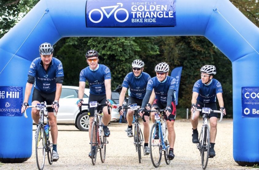 Cyclists ride under a blue inflatable arch labeled "Bidwells Golden Triangle Bike Ride" in an outdoor setting with trees and parked cars.